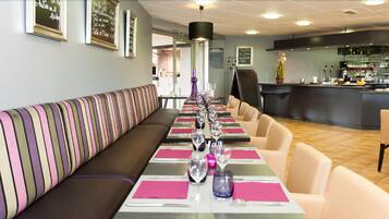 A restaurant with a long dining table set with pink napkins, silverware, and wine glasses. There is a striped bench along the wall and a bar area in the background.