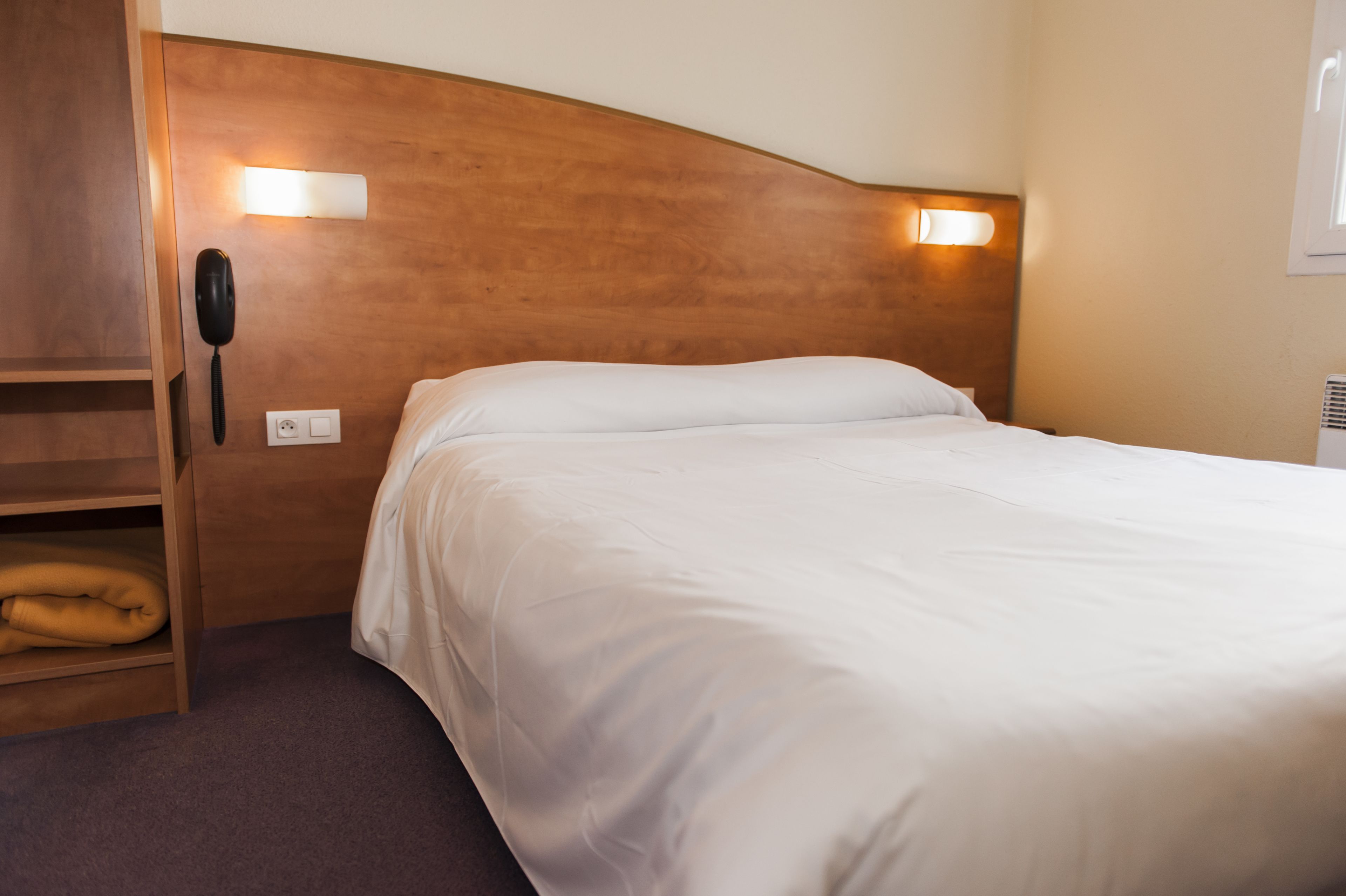 Une chambre d’hôtel avec une tête de lit en bois, un lit blanc, un téléphone sur la table de chevet et une étagère sur laquelle est pliée une couverture.