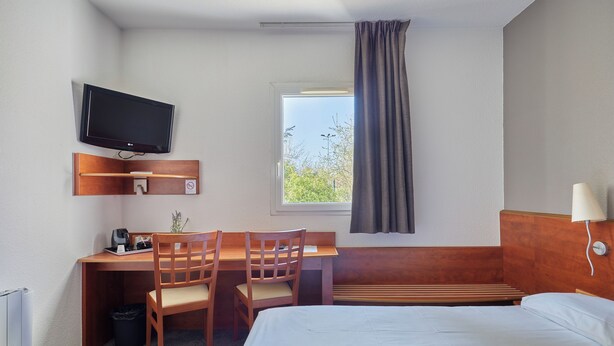 A hotel room with a bed, desk, chairs, a television, a window with curtains, and a lamp. - Citotel Armony Hotel (Bourg-Achard)