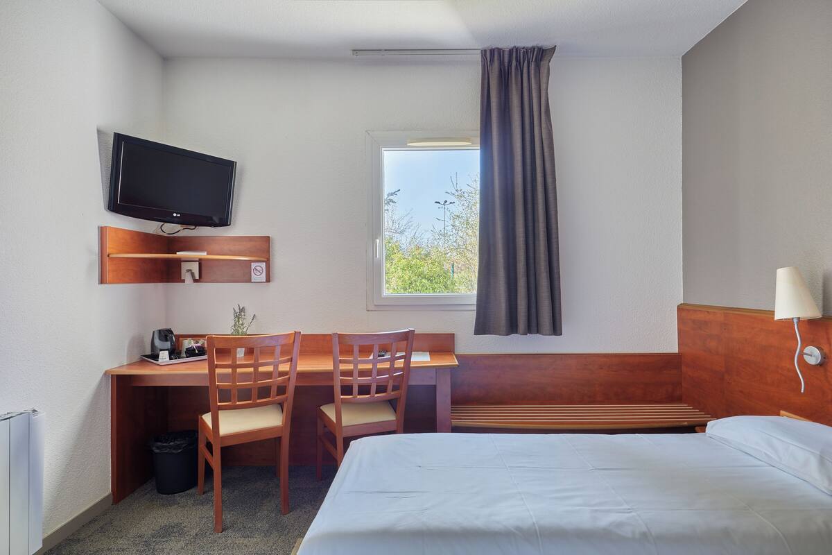 Ein Hotelzimmer mit Bett, Schreibtisch, Stühlen, einem Fernseher, einem Fenster mit Vorhängen und einer Lampe.
