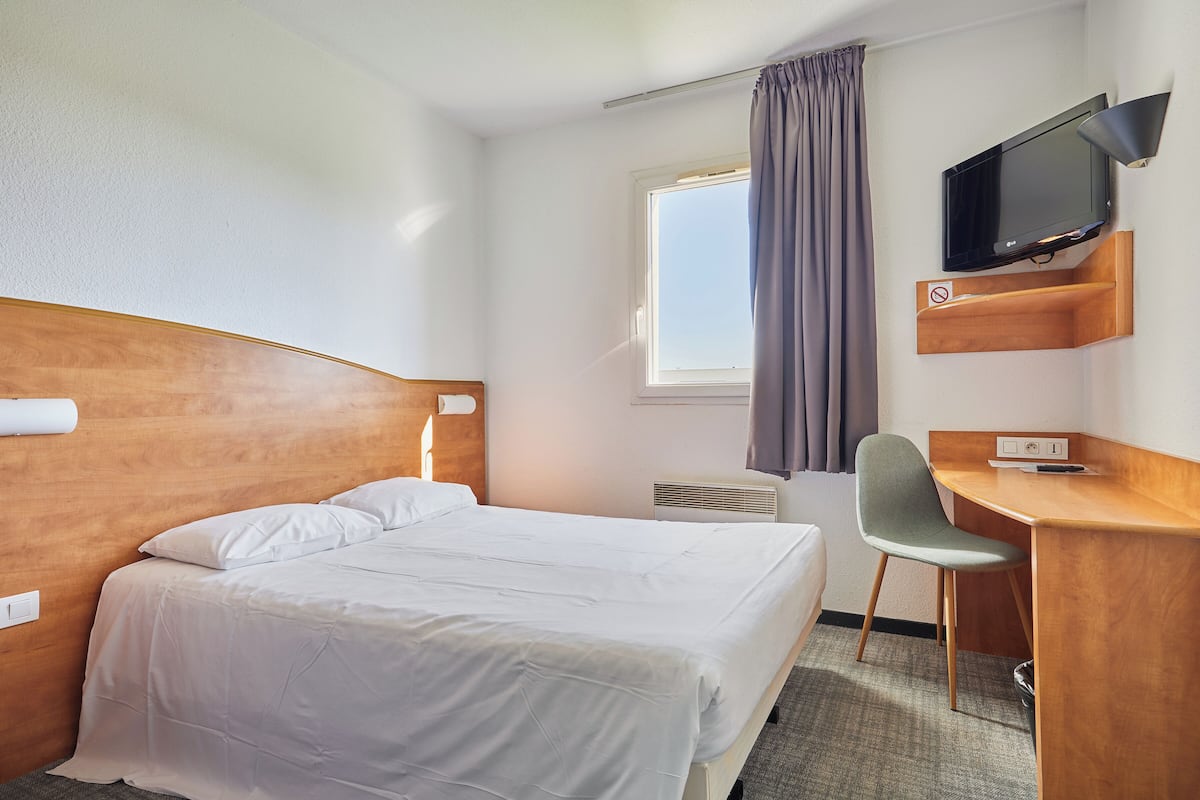 Ein Hotelzimmer mit Bett, Schreibtisch, Sessel, Fernseher und einem fensterbepflanzten Fenster.