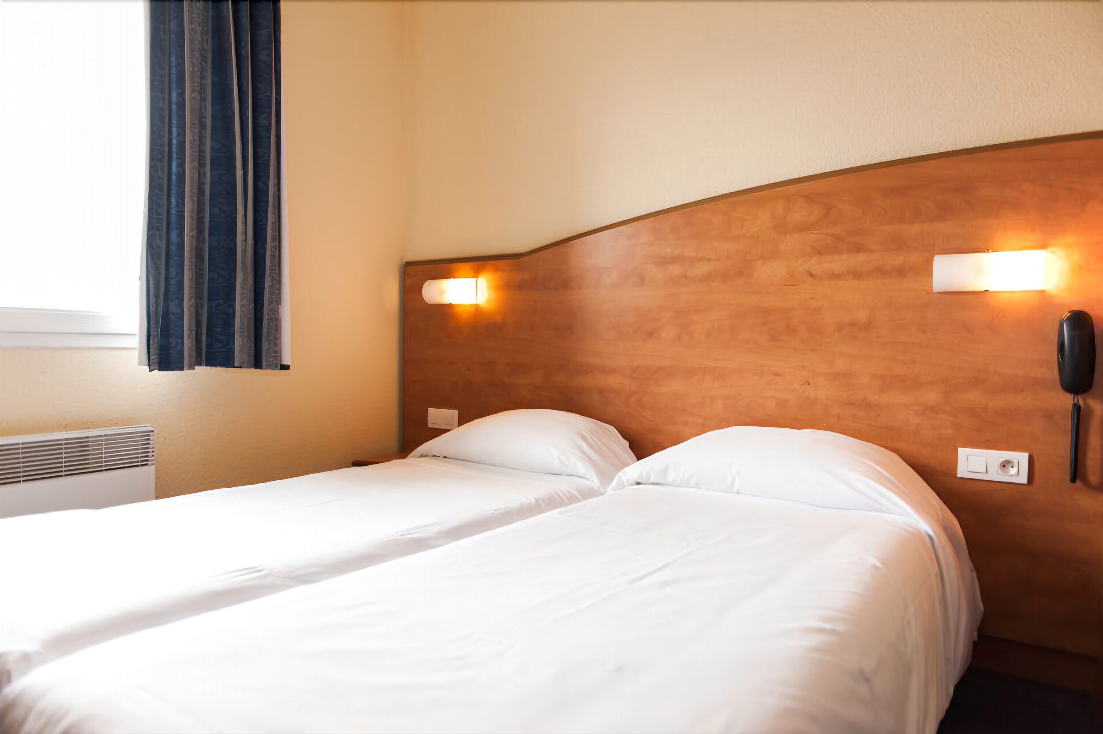 A hotel room with two single beds, a wooden headboard, bedside lamps, a phone, and a window with curtains.