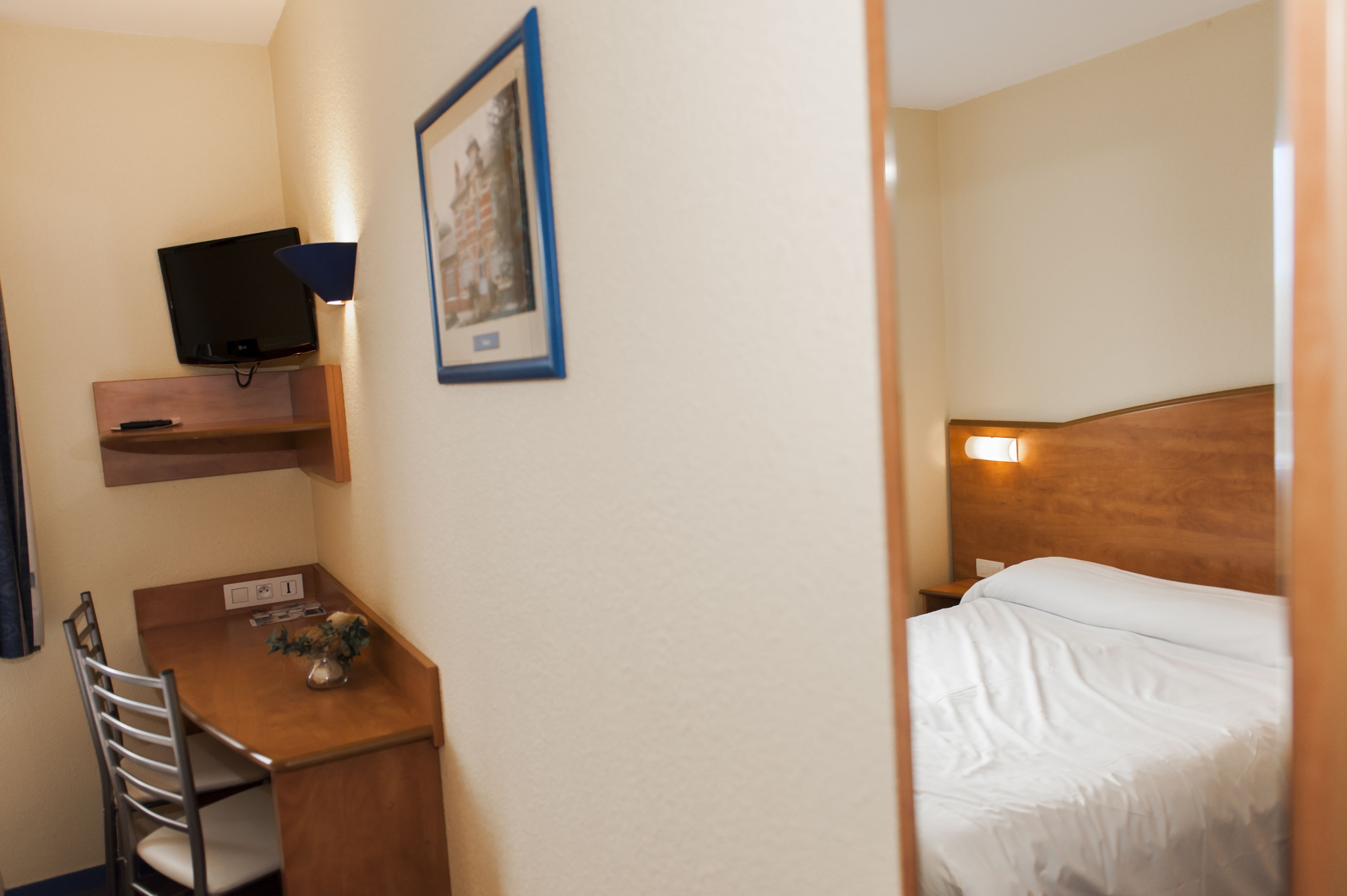 A hotel room with a bed, a desk with a chair, a television mounted on the wall, and a framed picture.