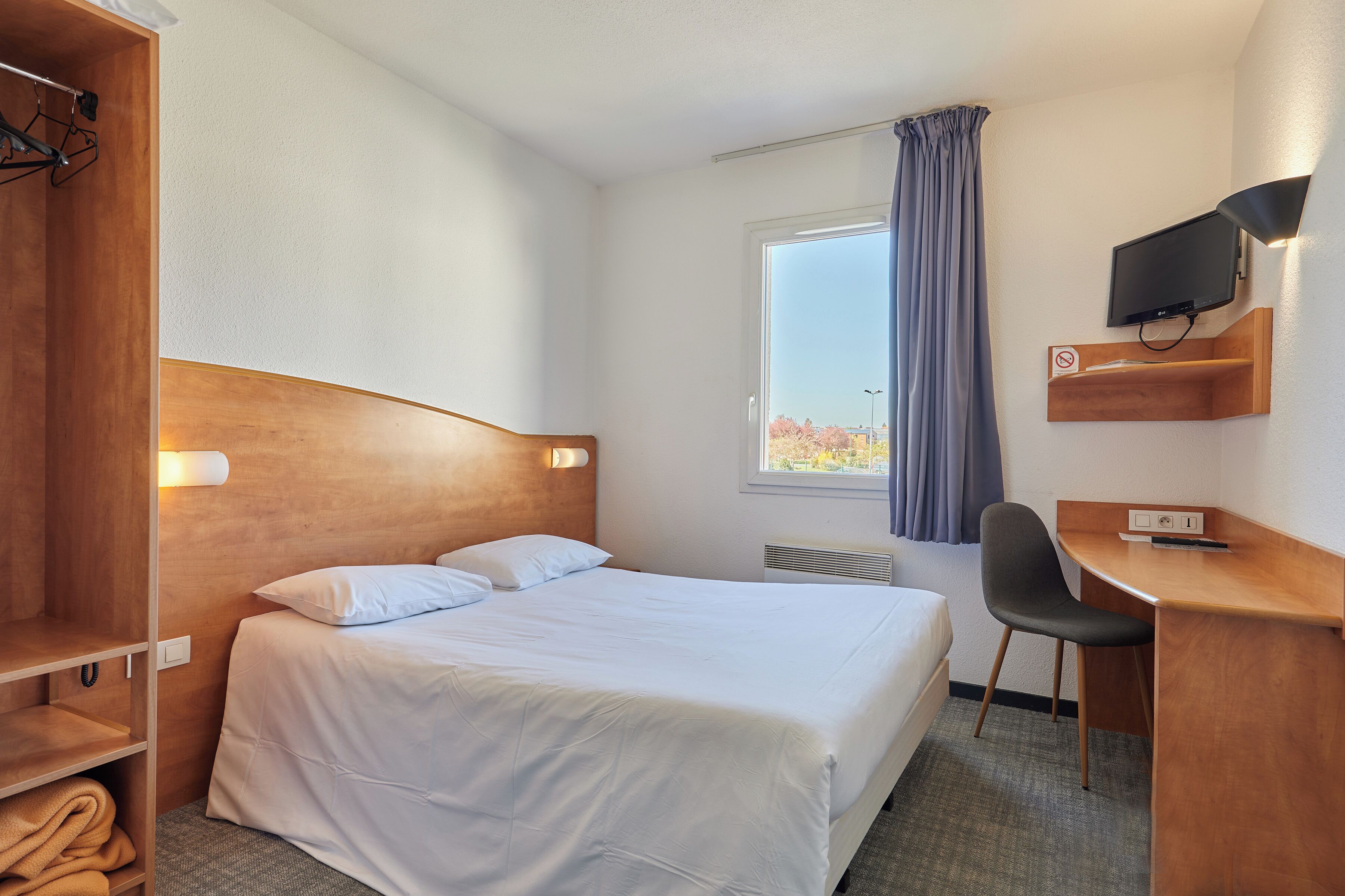 Une chambre d’hôtel équipée d’un lit, d’un bureau, d’une chaise, d’une télévision, d’une armoire et d’une fenêtre avec des rideaux.