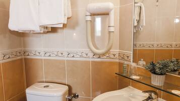 A bathroom with a toilet, a sink, a glass shelf with toiletries, and a hairdryer on the wall.