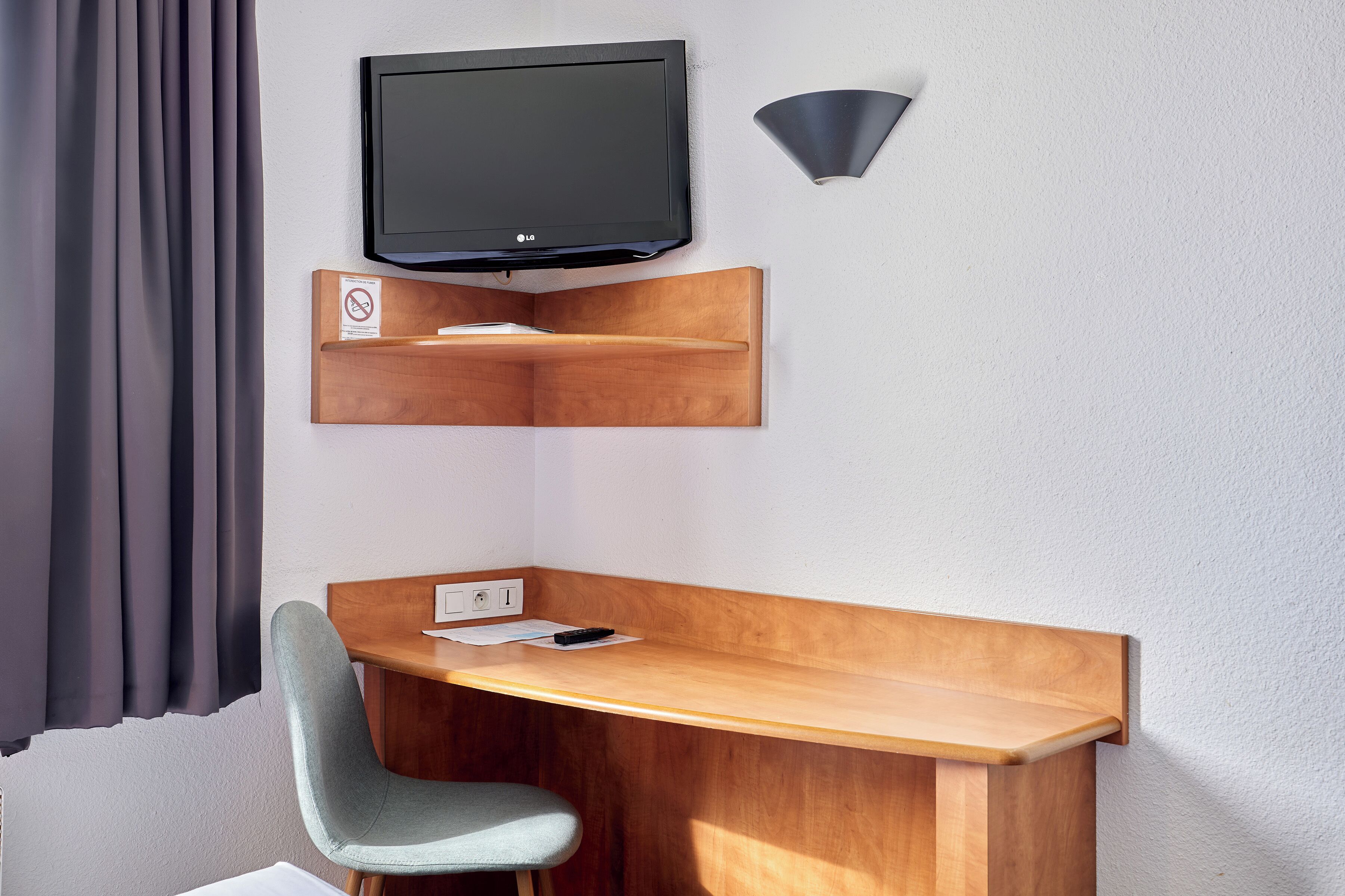 Habitación de hotel moderna con escritorio de madera, silla gris, televisor montado y lámpara fijada a la pared.