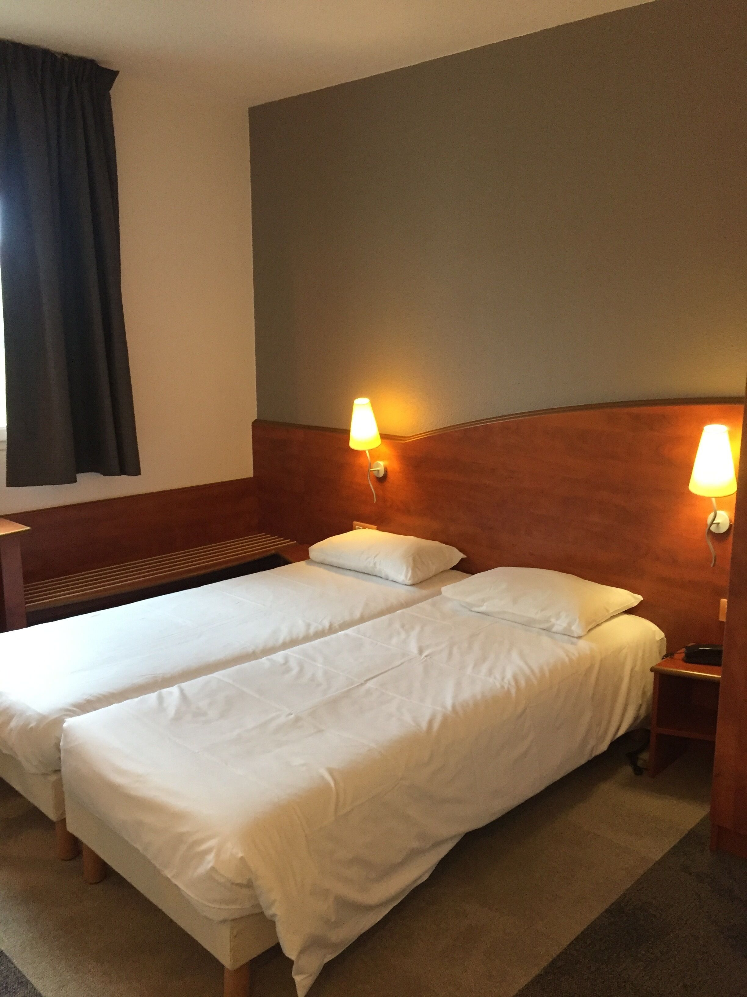 A hotel room with two beds, a wooden headboard, a nightstand, and a lamp.