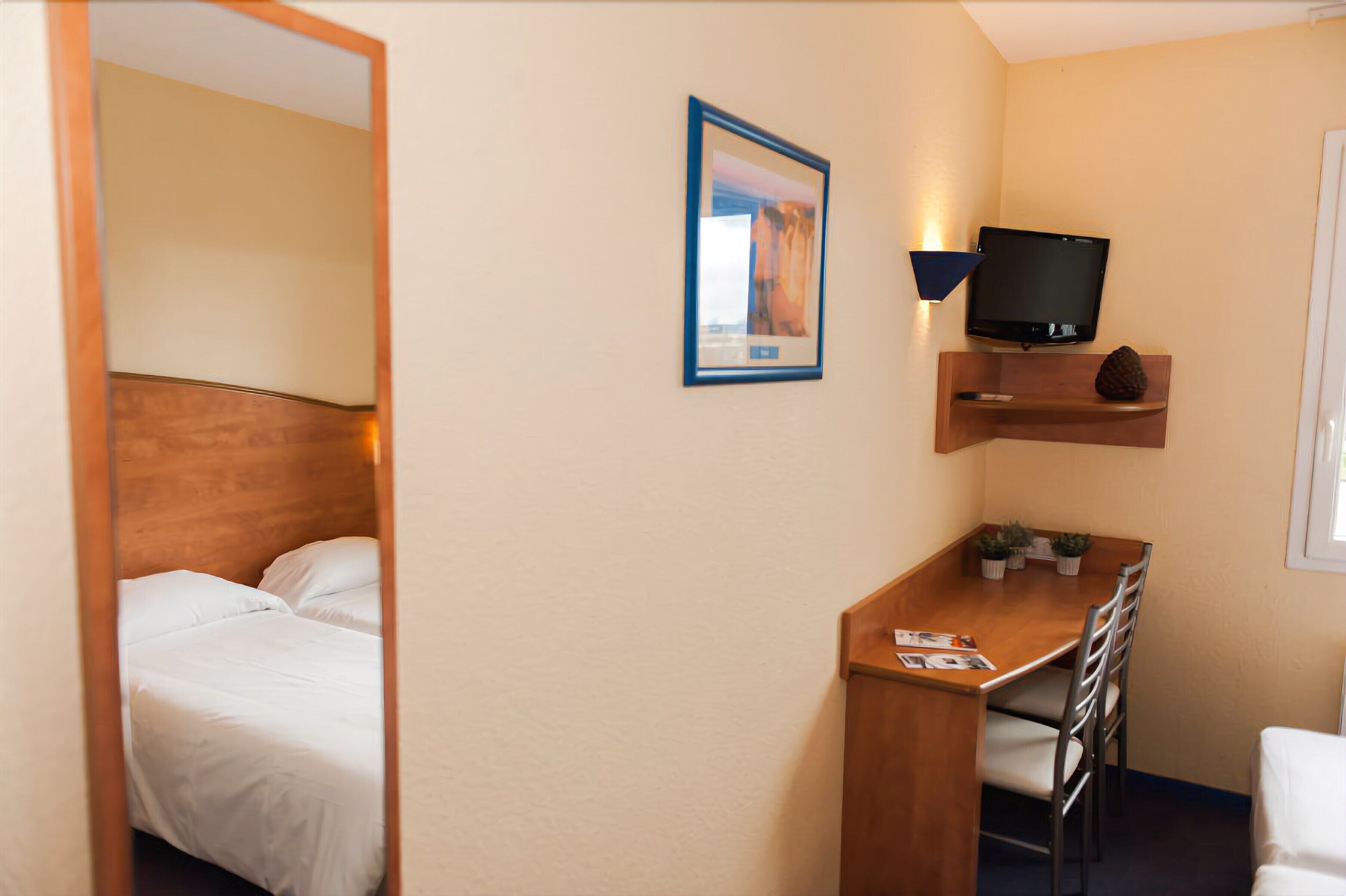 Une chambre d’hôtel avec un lit, un bureau avec des chaises, une télévision et une petite étagère avec une plante.
