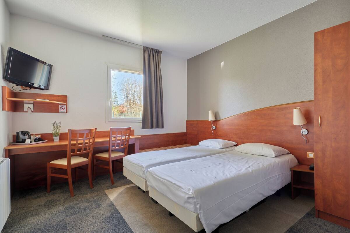 Ein Hotelzimmer mit Schreibtisch, zwei Betten, einem Fernseher, einem Fenster mit Vorhängen und einem Holz-Kopfstück.