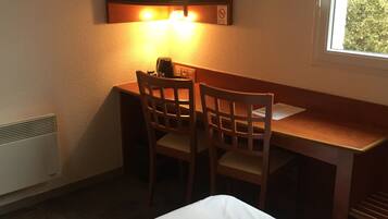A hotel room with a bed, a desk with two chairs, a television mounted on the wall, and a window with a view of greenery.