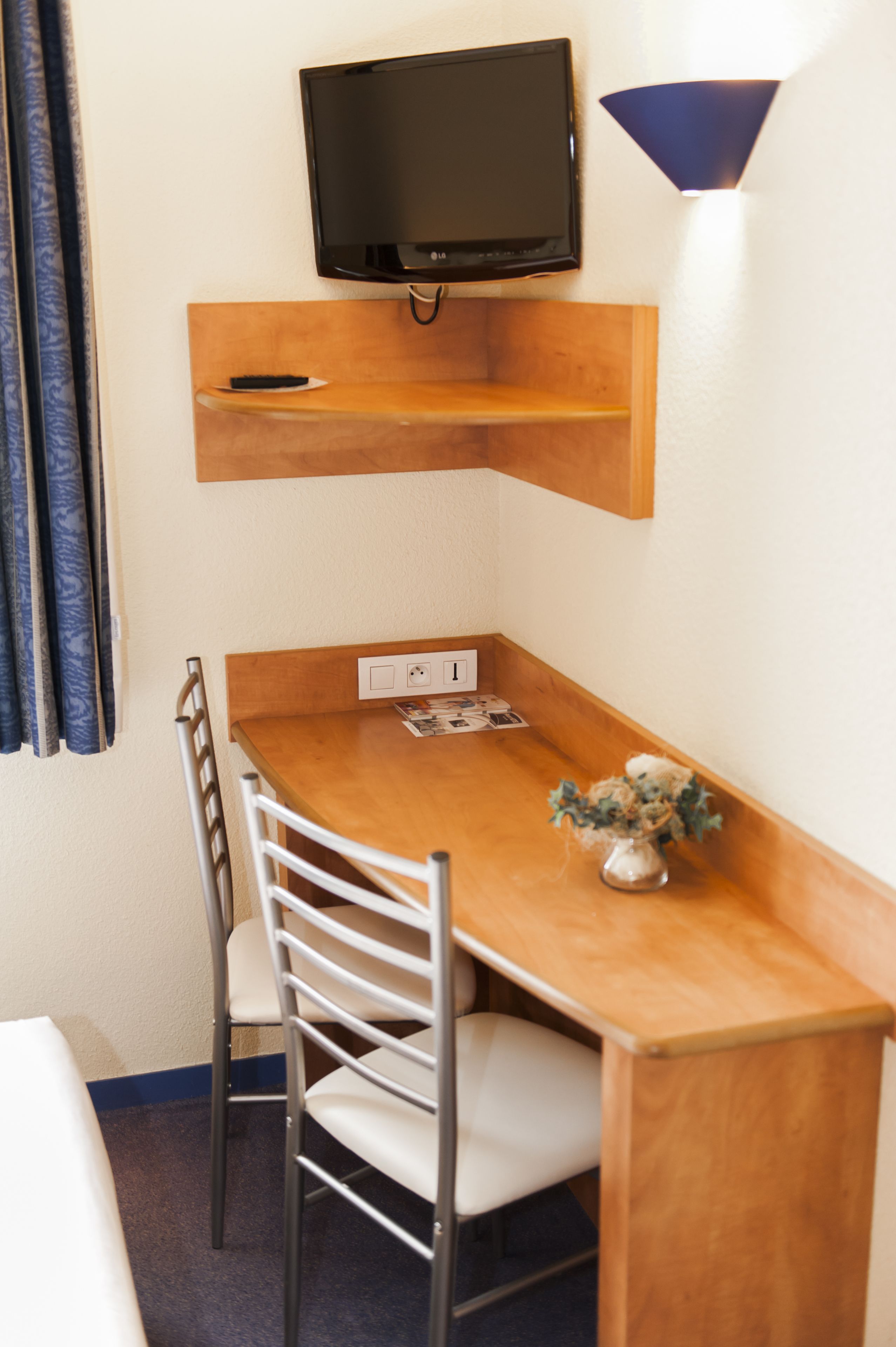 Una habitación pequeña y ordenada con una mesa de madera, dos sillas, un televisor de pantalla plana montado en la pared y una pequeña estantería sobre la mesa.