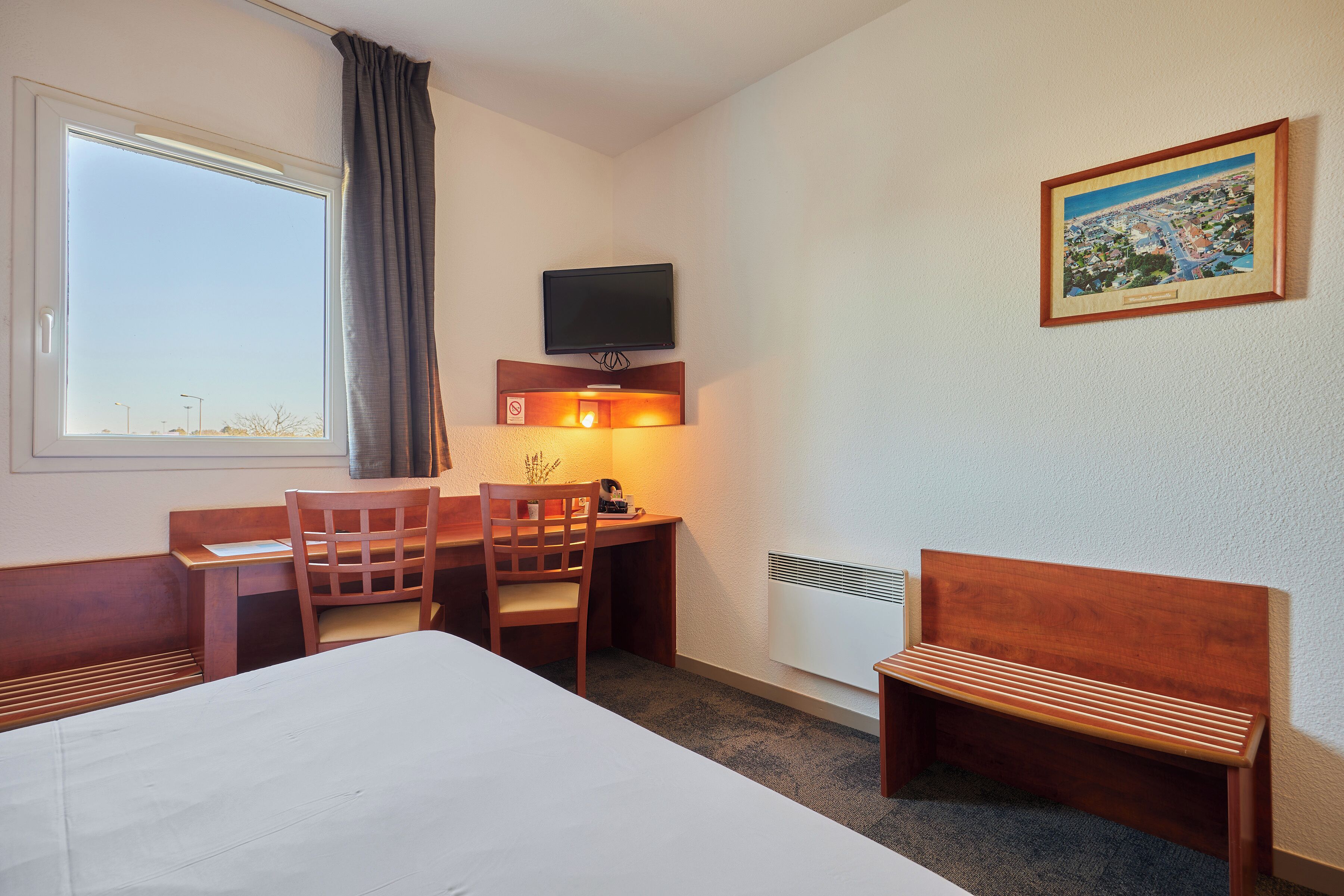 Ein Hotelzimmer mit Bett, Schreibtisch mit Stuhl, Fernseher, Heizkörper und einem Fenster mit Vorhängen.