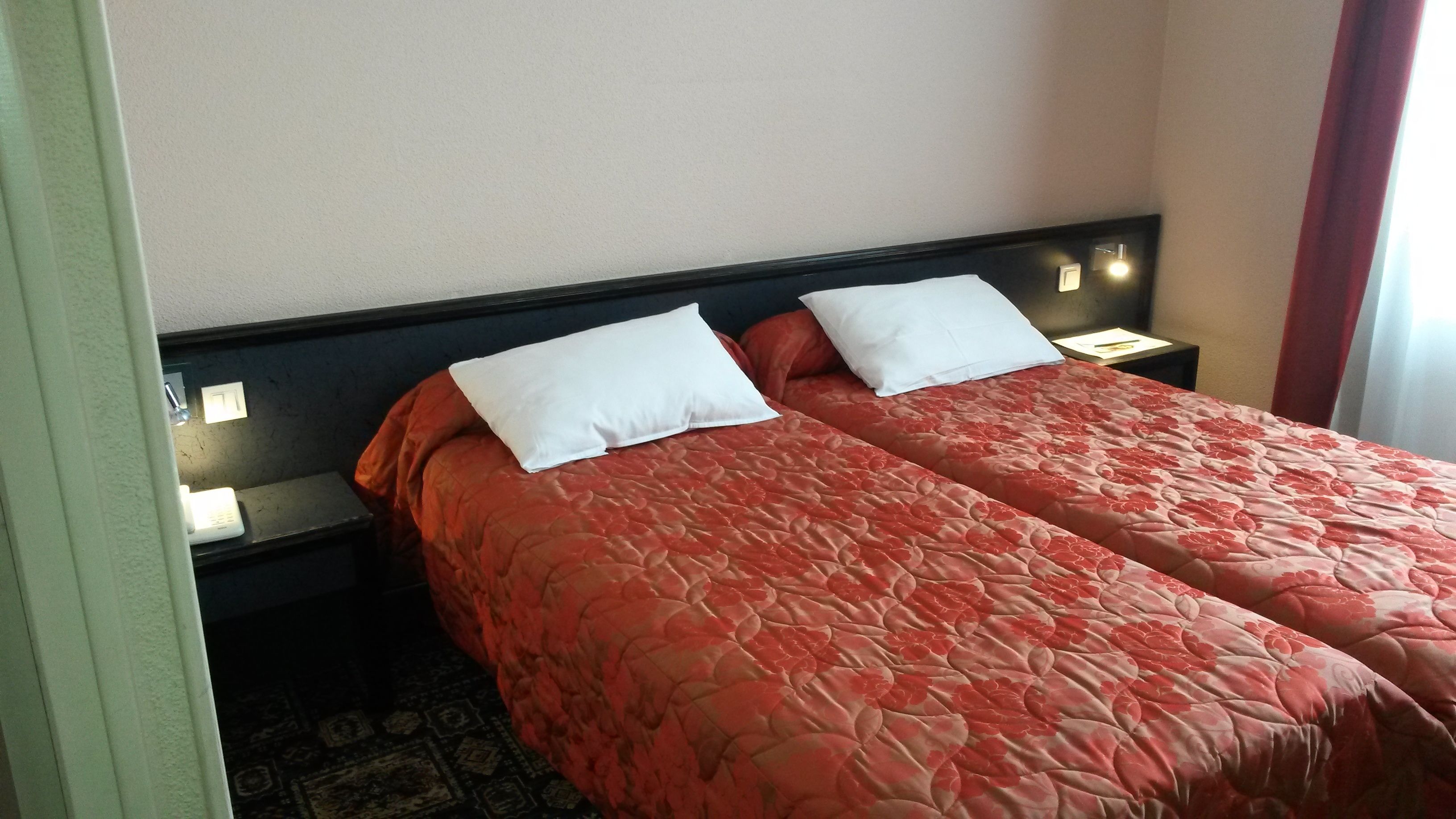 A hotel room with two beds, each with a red floral bedspread and white pillows. There are bedside tables with lamps and a small clock. The room has a simple, clean design with a carpeted floor.