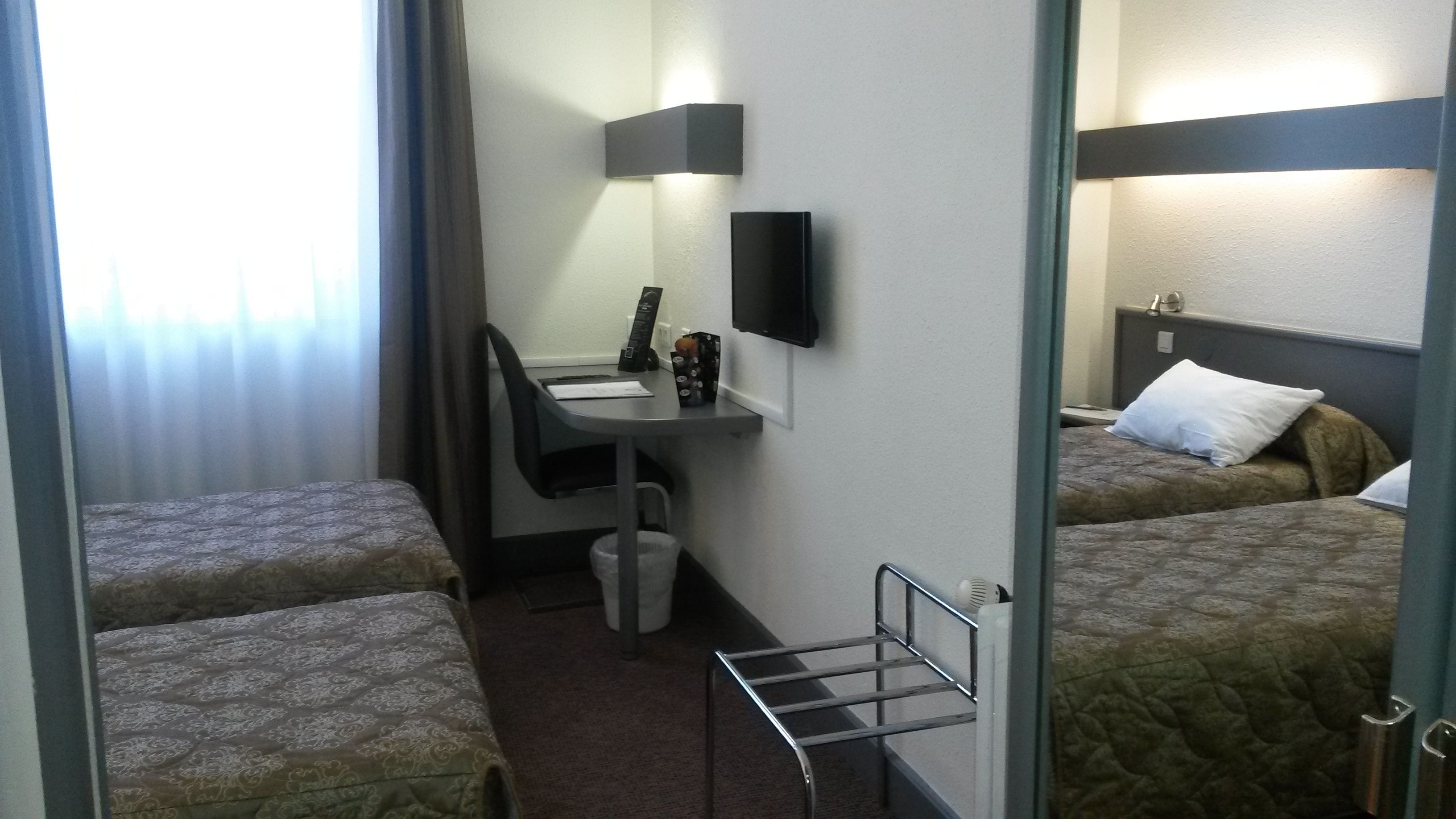 A hotel room with two single beds, a desk with a chair, a small table, a TV, and a window with curtains.