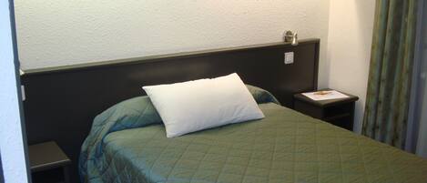 Une chambre d’hôtel avec un lit simple, une table de chevet, une lampe et un rideau.
