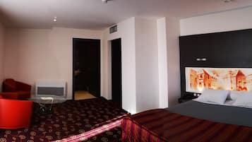 A hotel room with a bed, a red armchair, a small table, and a wall-mounted TV.