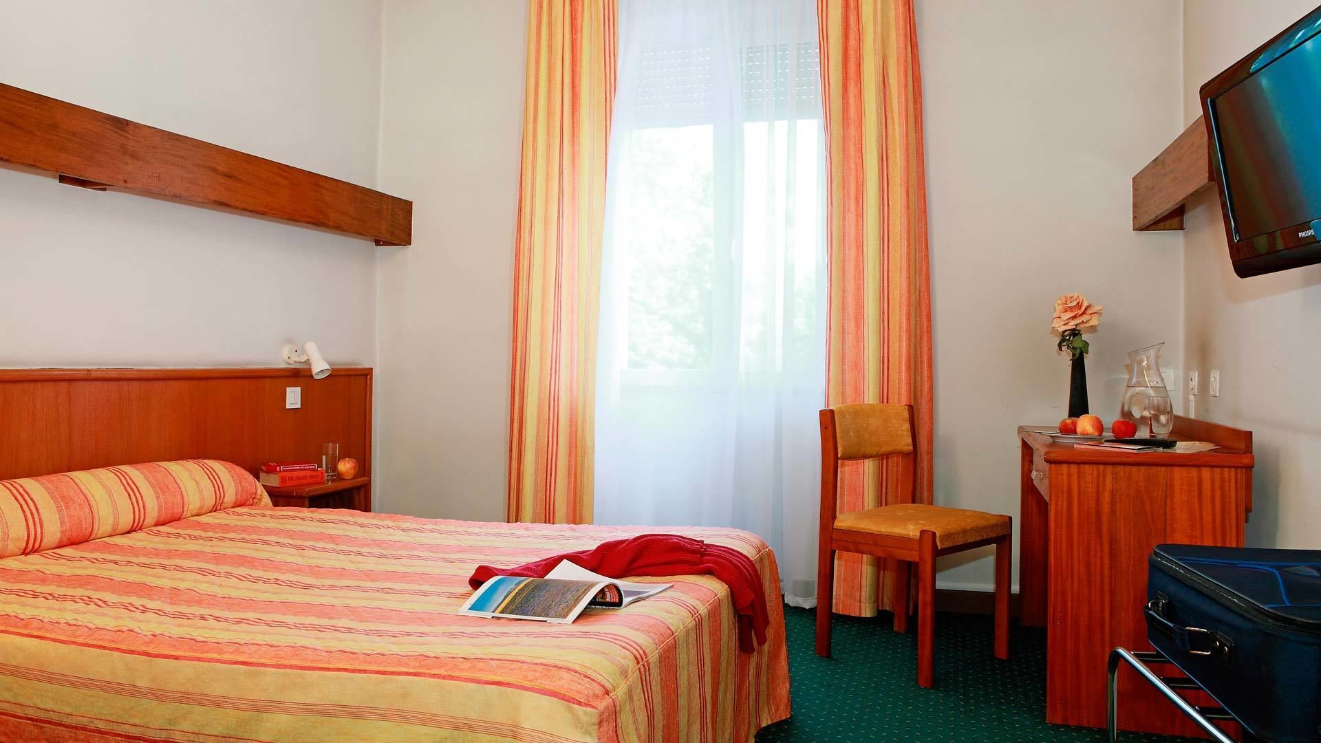 Habitación de hotel con cama, escritorio, silla, televisor y ventana con cortinas.