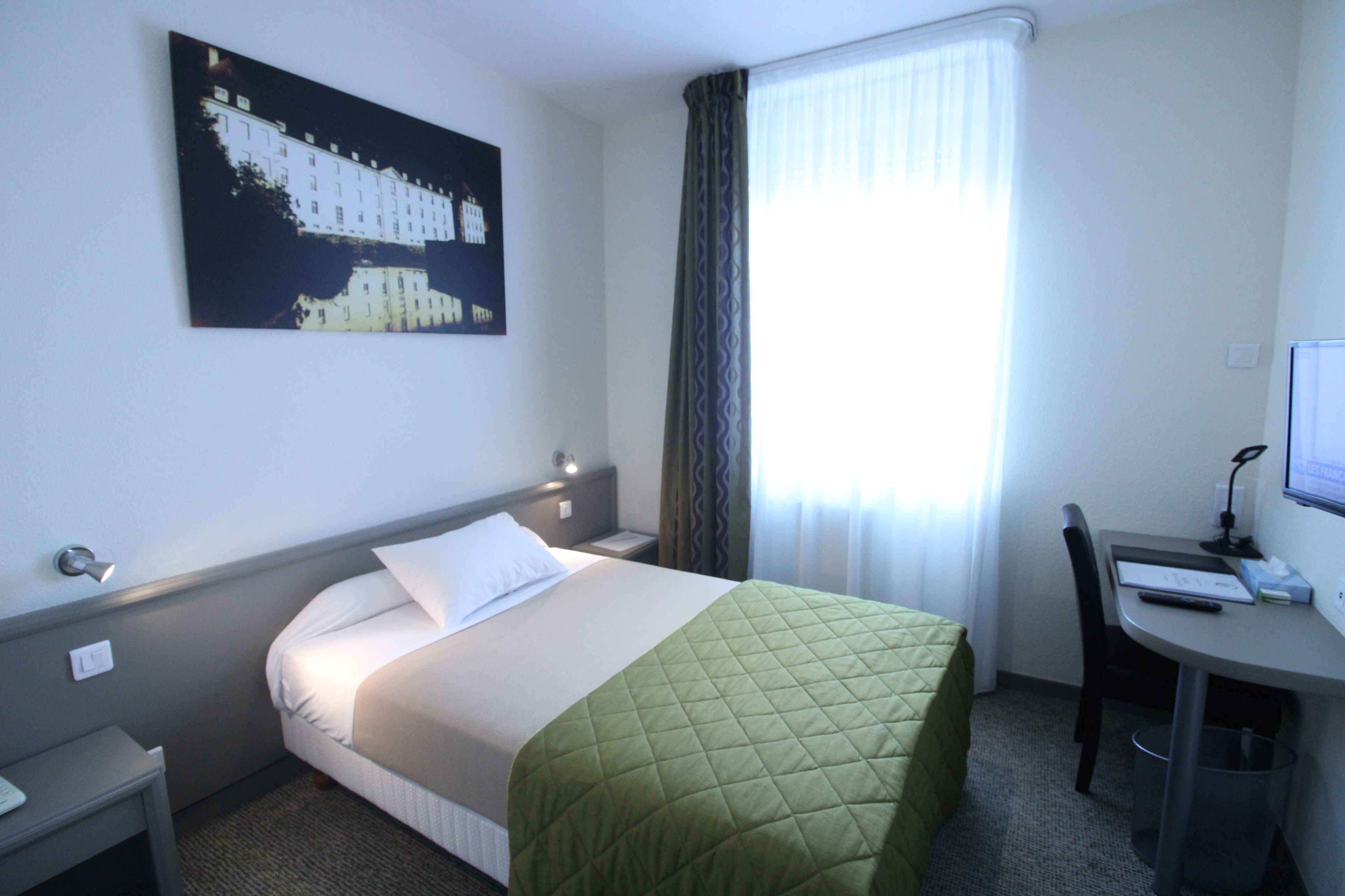 A hotel room with a bed, a desk, a chair, a computer, and a window with curtains.