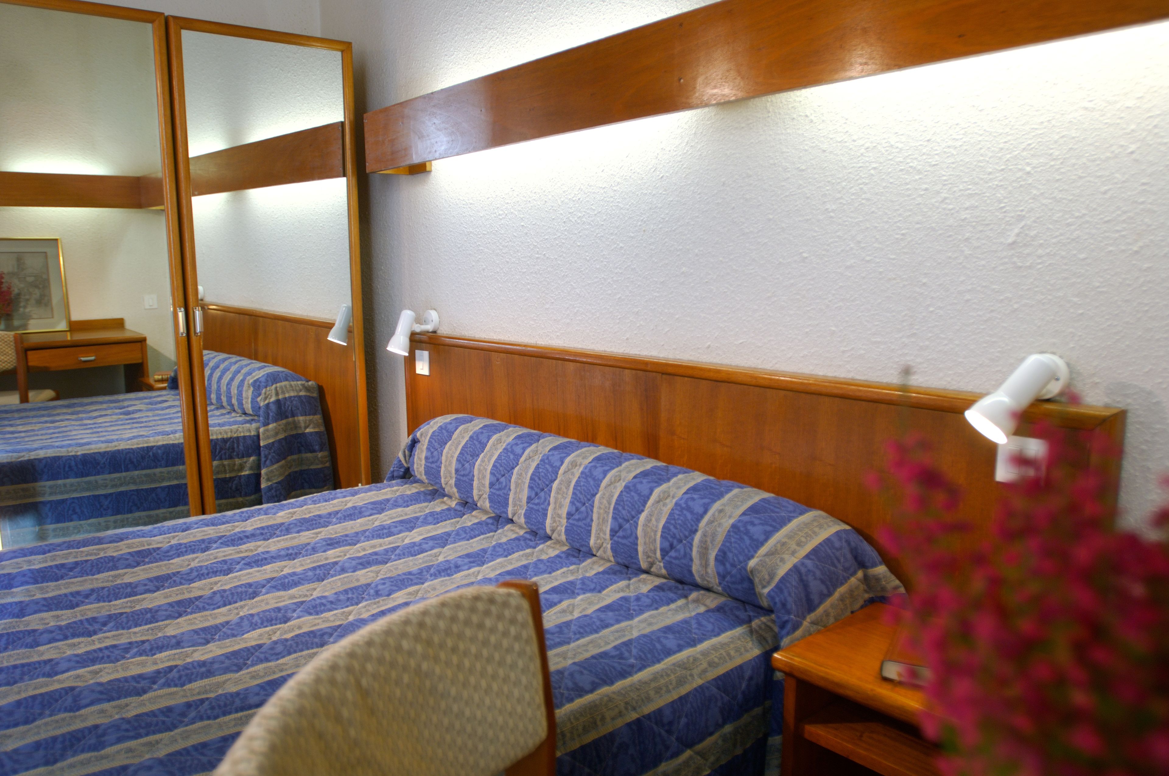 A hotel room with two beds, wooden headboards, a bedside table with a lamp, and a chair.