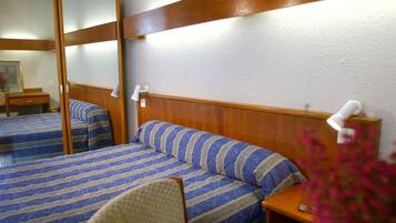 A hotel room with two beds, wooden headboards, a bedside table with a lamp, and a chair.