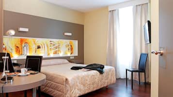 A hotel room with a bed, a desk with a laptop, a chair, a TV mounted on the wall, and a large window with curtains.