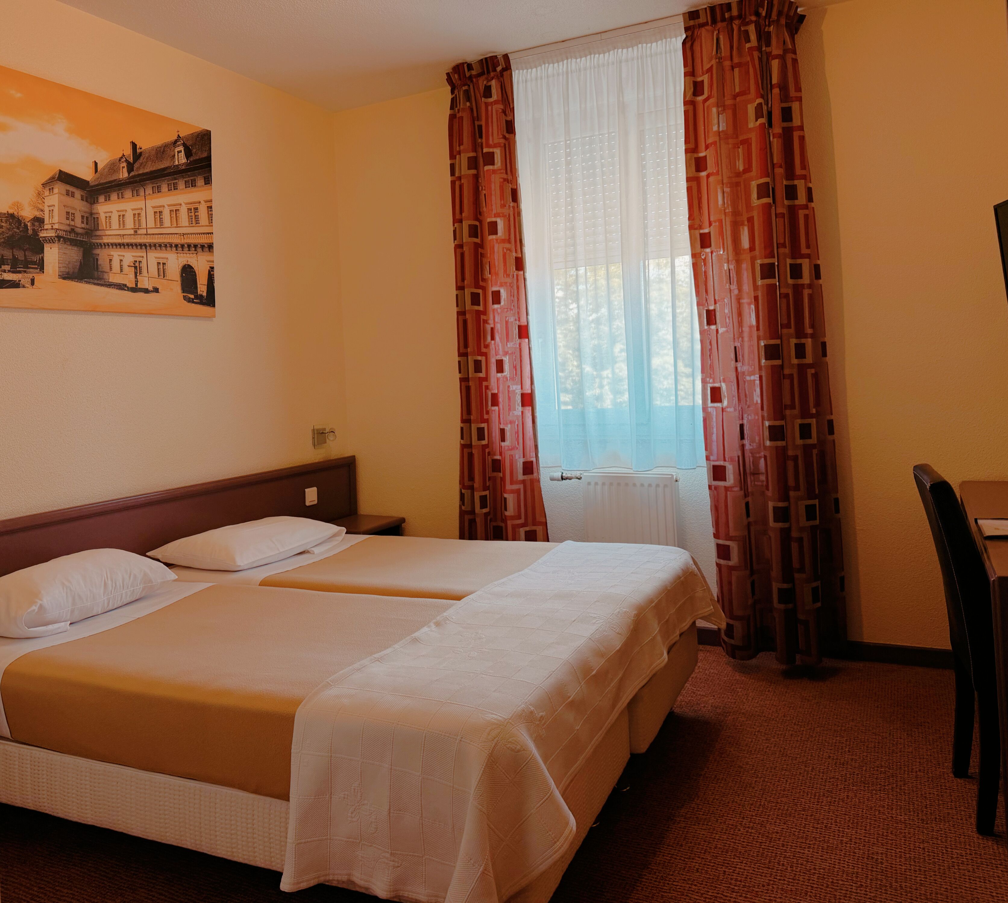 Habitación de hotel con dos camas, un escritorio, una silla, un ventanal con cortinas y un cuadro en la pared.