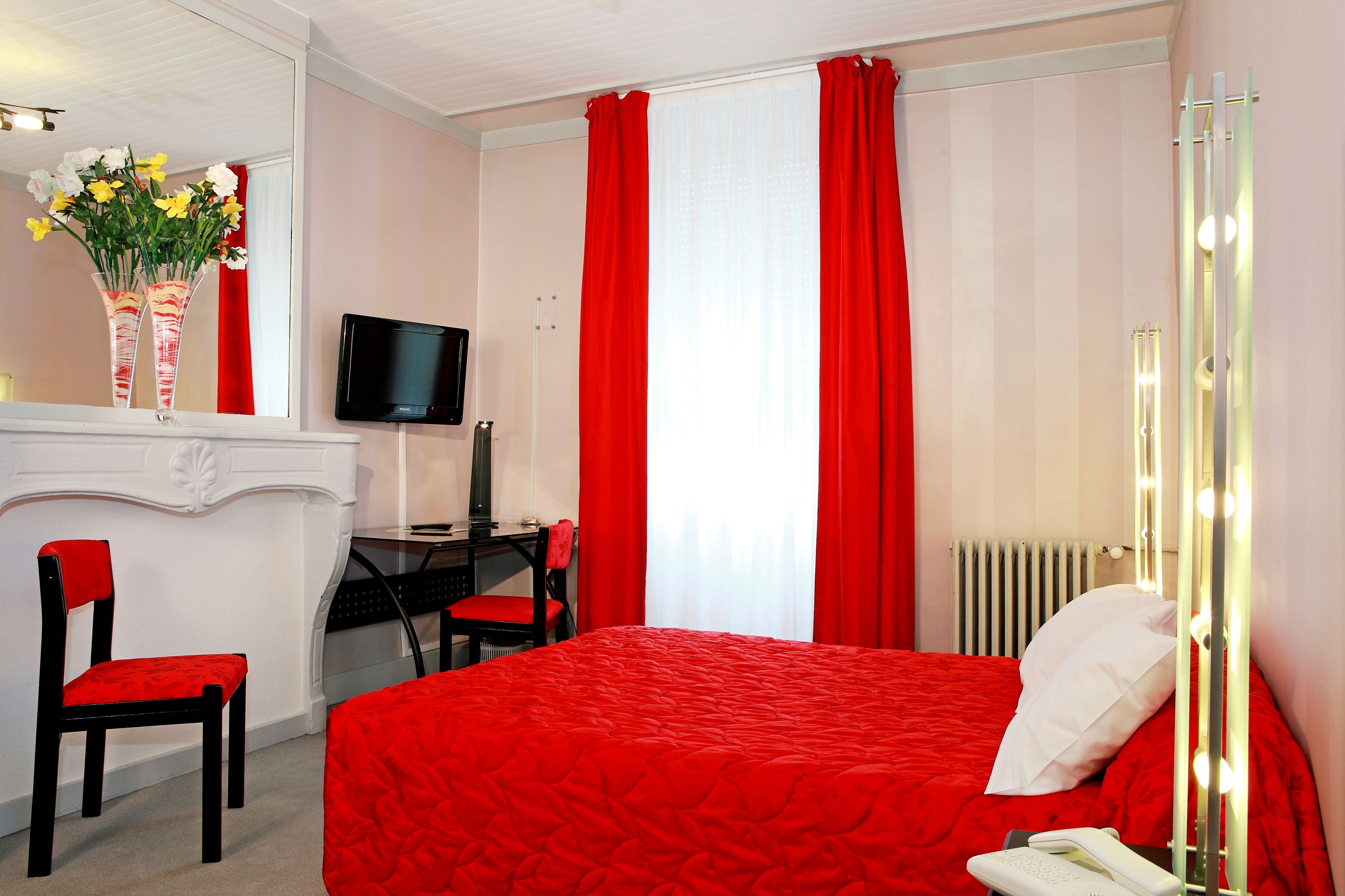 A hotel room with a red bedspread, a desk with a computer, a chair, a mirror, and a TV.