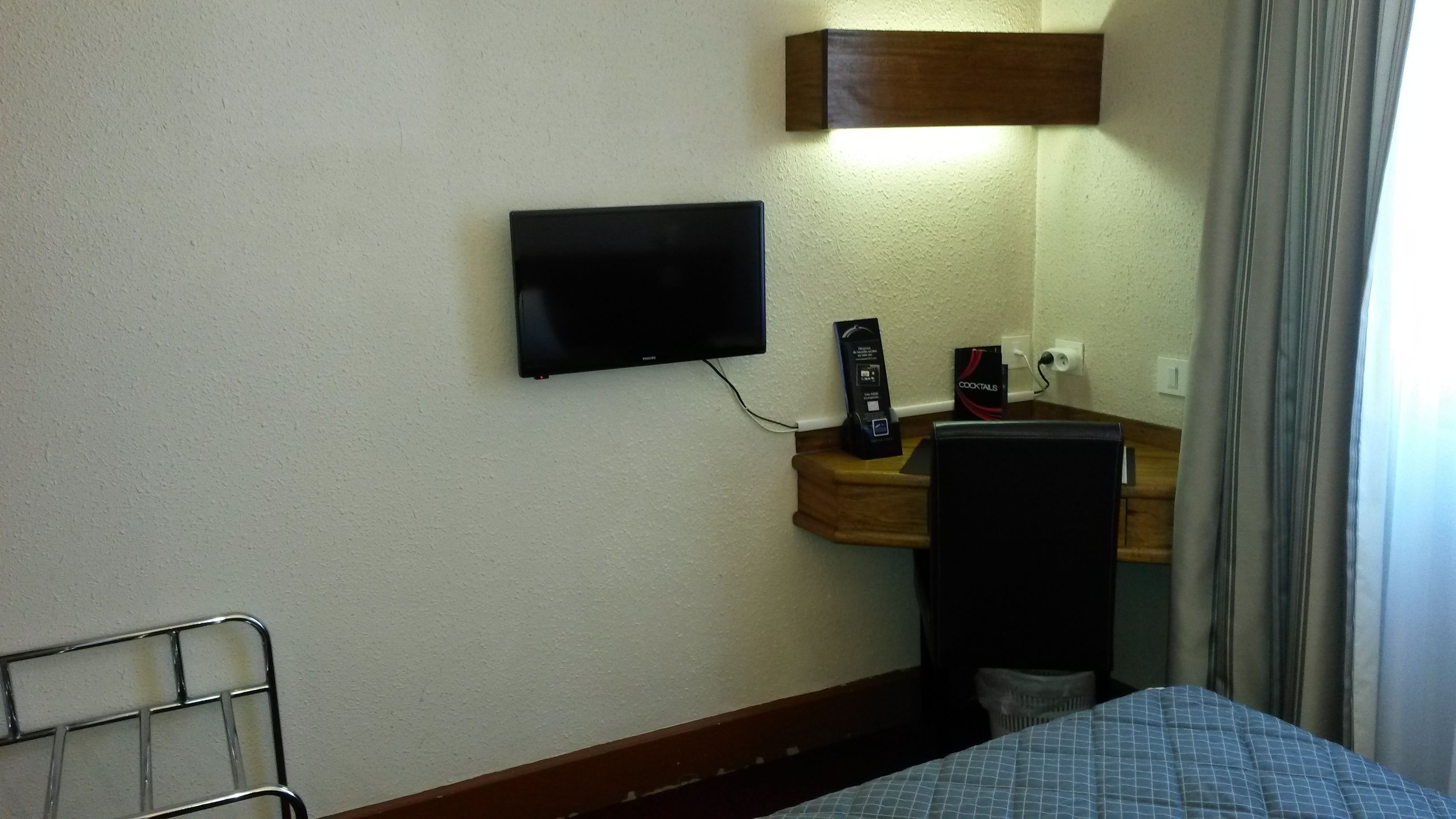 A hotel room with a bed, a TV mounted on the wall, a small wooden side table, and a mirror.