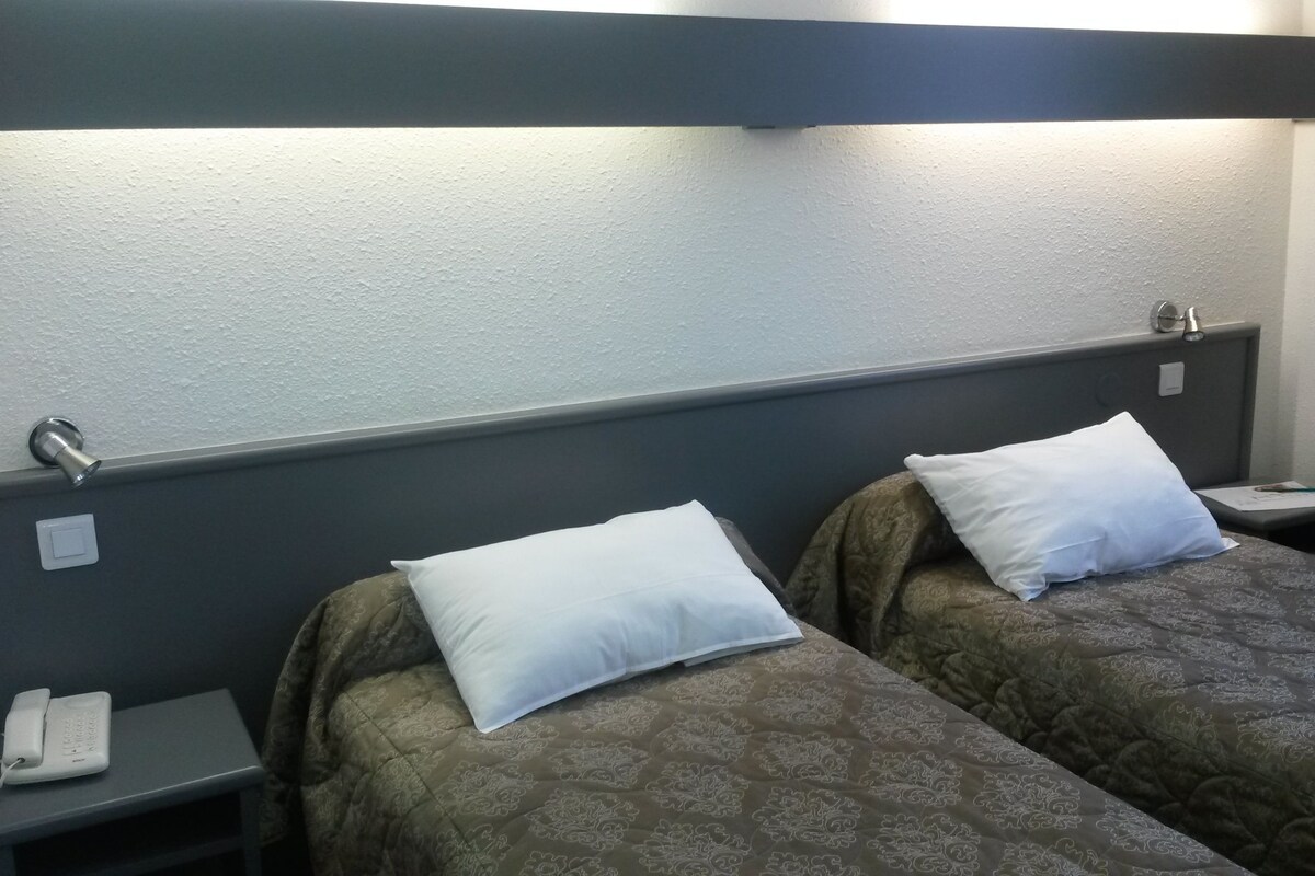 A hotel room with two single beds, each with a pillow and a bedside table. There is a telephone on the left side of the room.