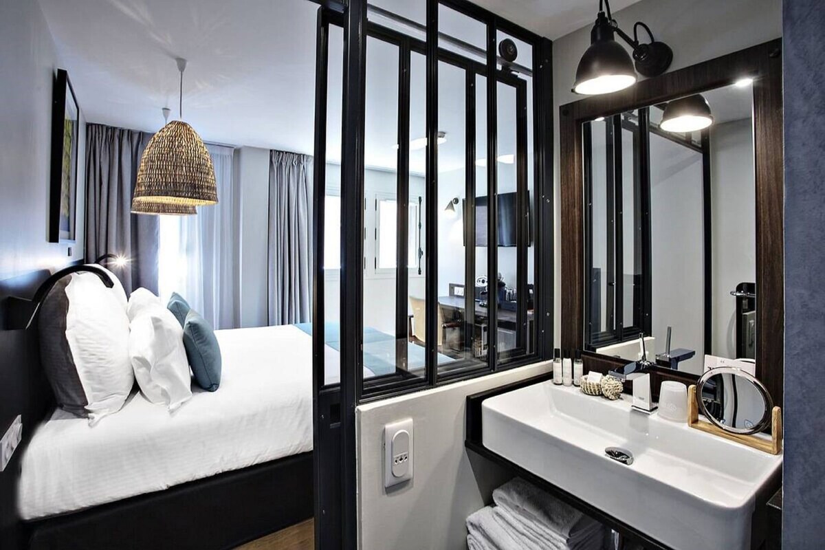 A hotel room with a bed, a large window, a bathroom with a sink and mirror, and a hanging light fixture.