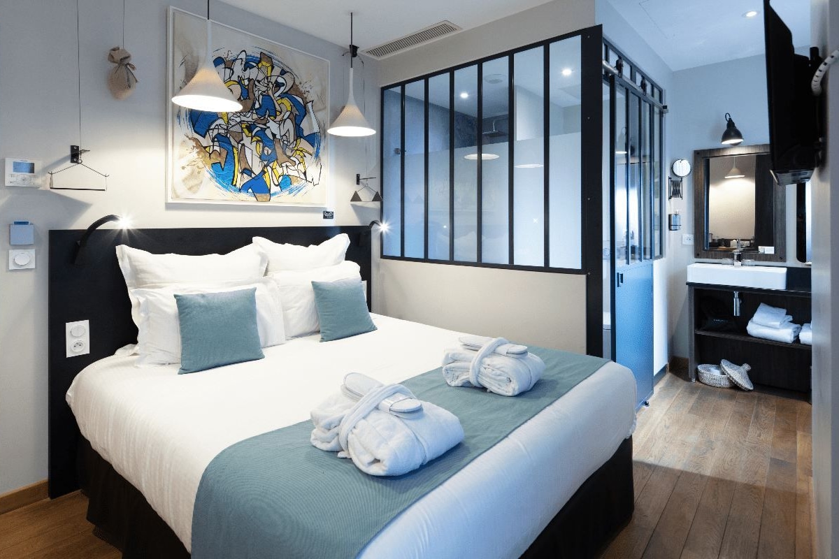 A modern hotel room with a large bed, a bathroom with a mirror and sink, and a large window with blinds.