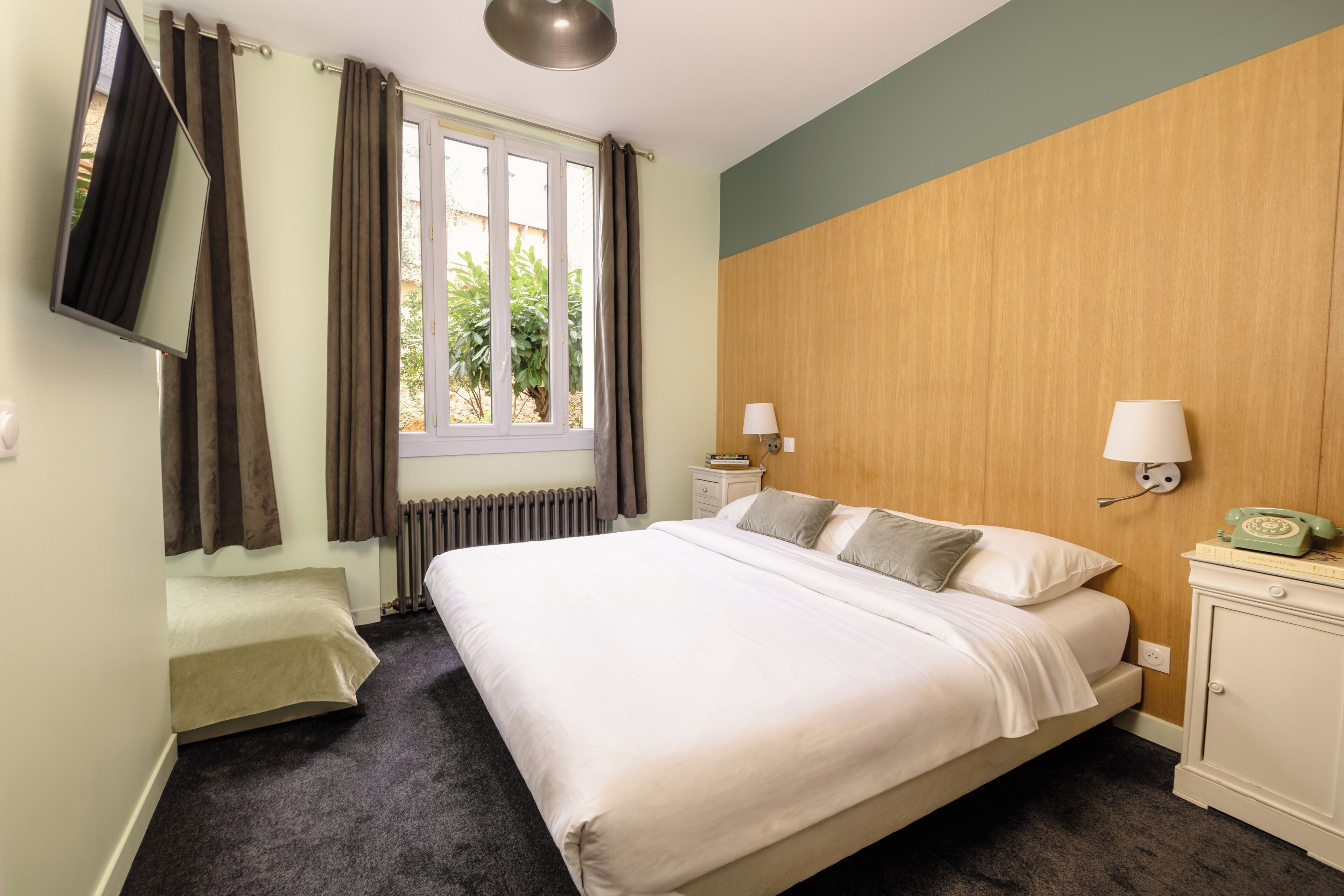 Habitación de hotel con cama, mesita de noche, lámpara, teléfono, ventana con cortinas y radiador.