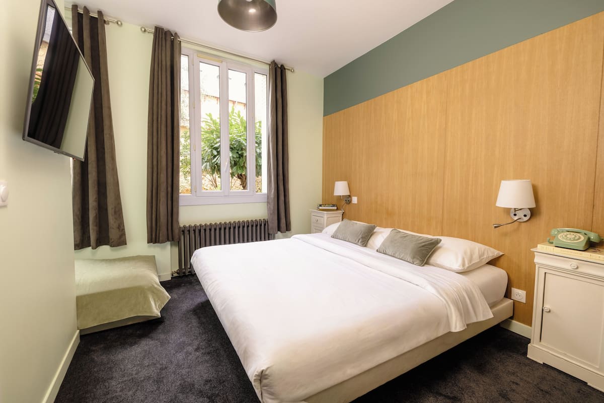 Habitación de hotel con cama, mesita de noche, lámpara, teléfono, ventana con cortinas y radiador.
