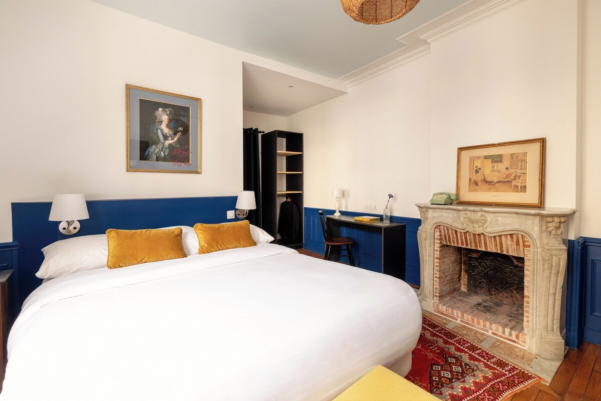 A hotel room with a large bed, a desk, a fireplace, and a bookshelf.
