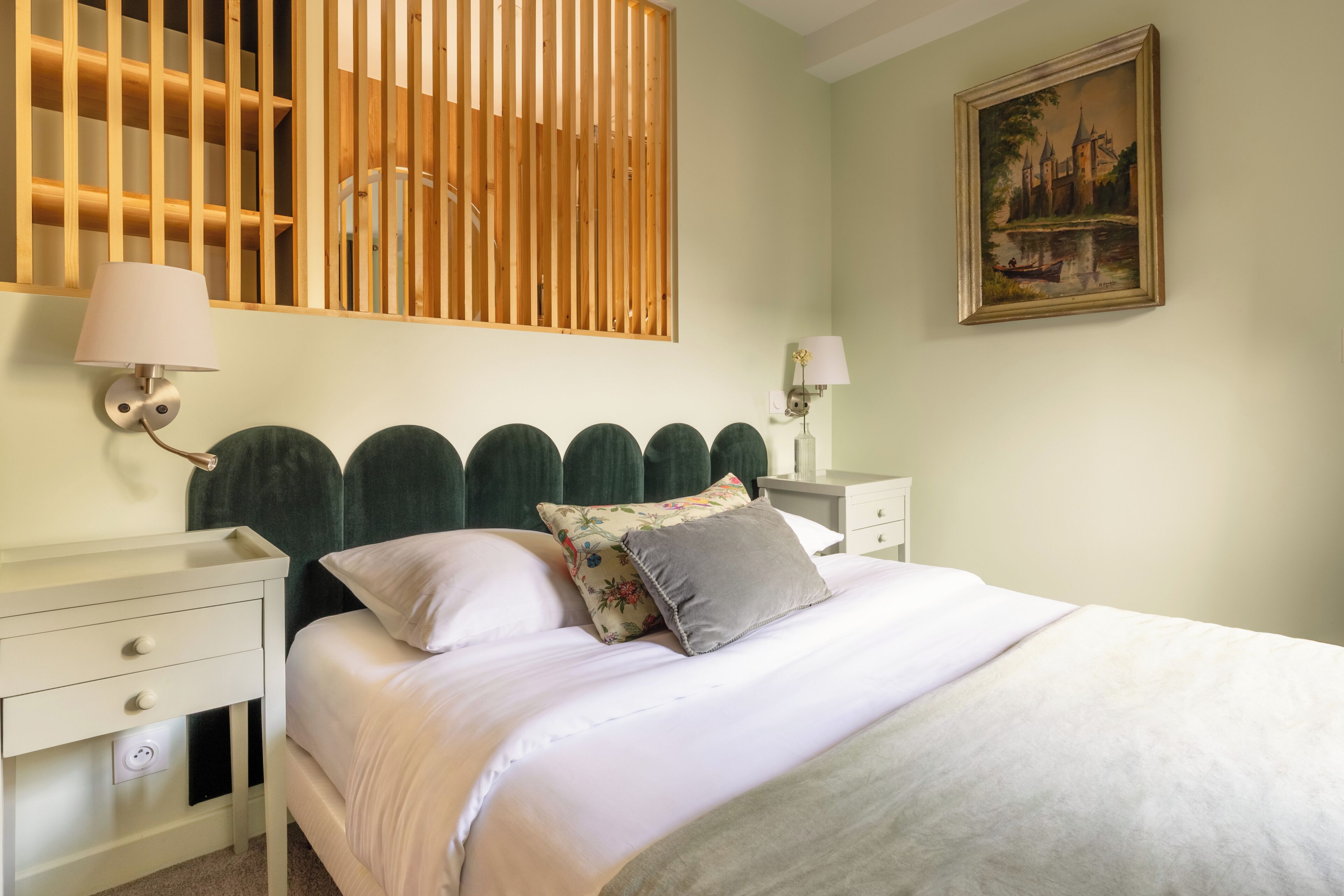 A bedroom with a bed, two bedside tables, a wall-mounted lamp, a painting, and a wooden slat partition.