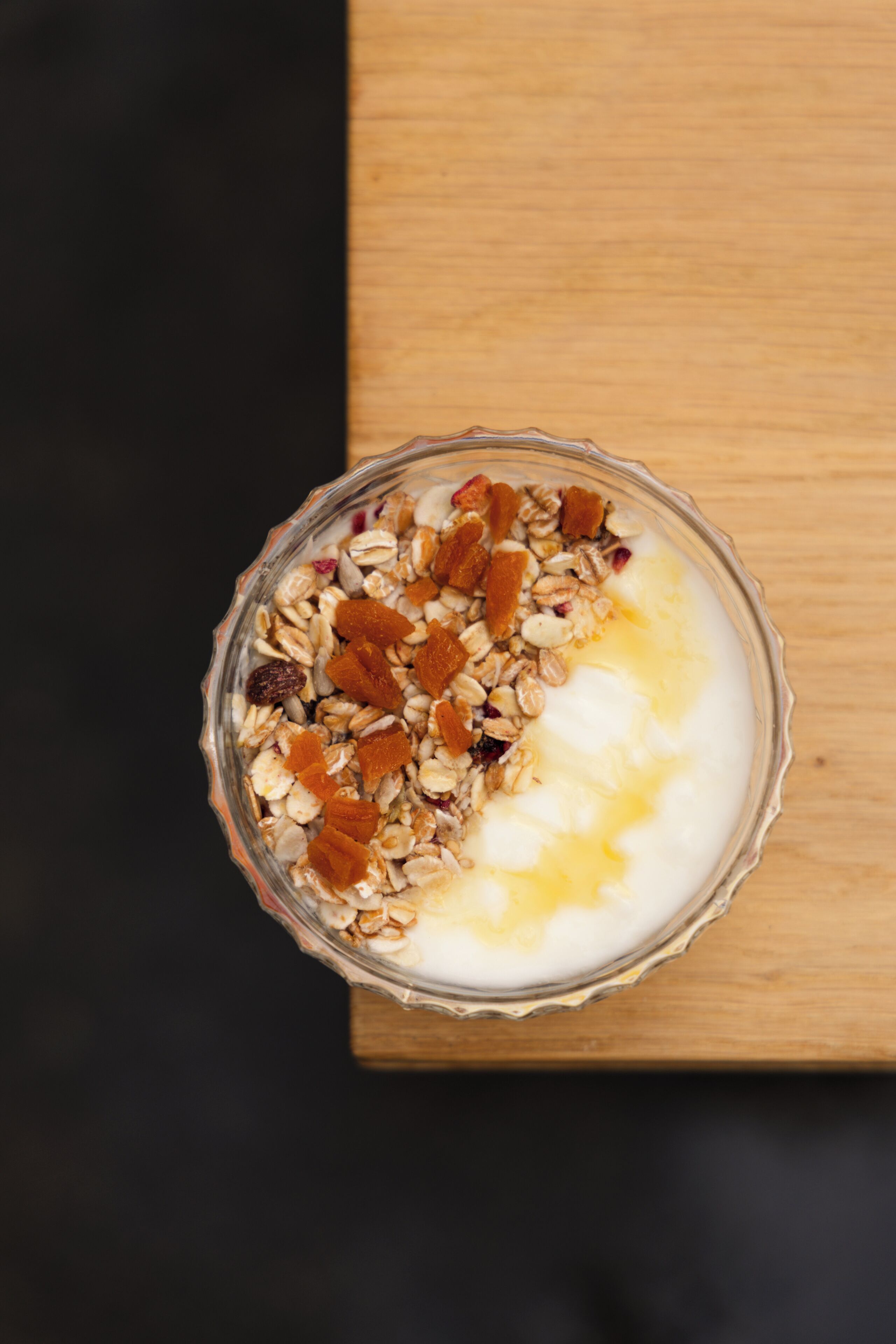 A glass bowl of yogurt with granola and dried fruit on a wooden surface.