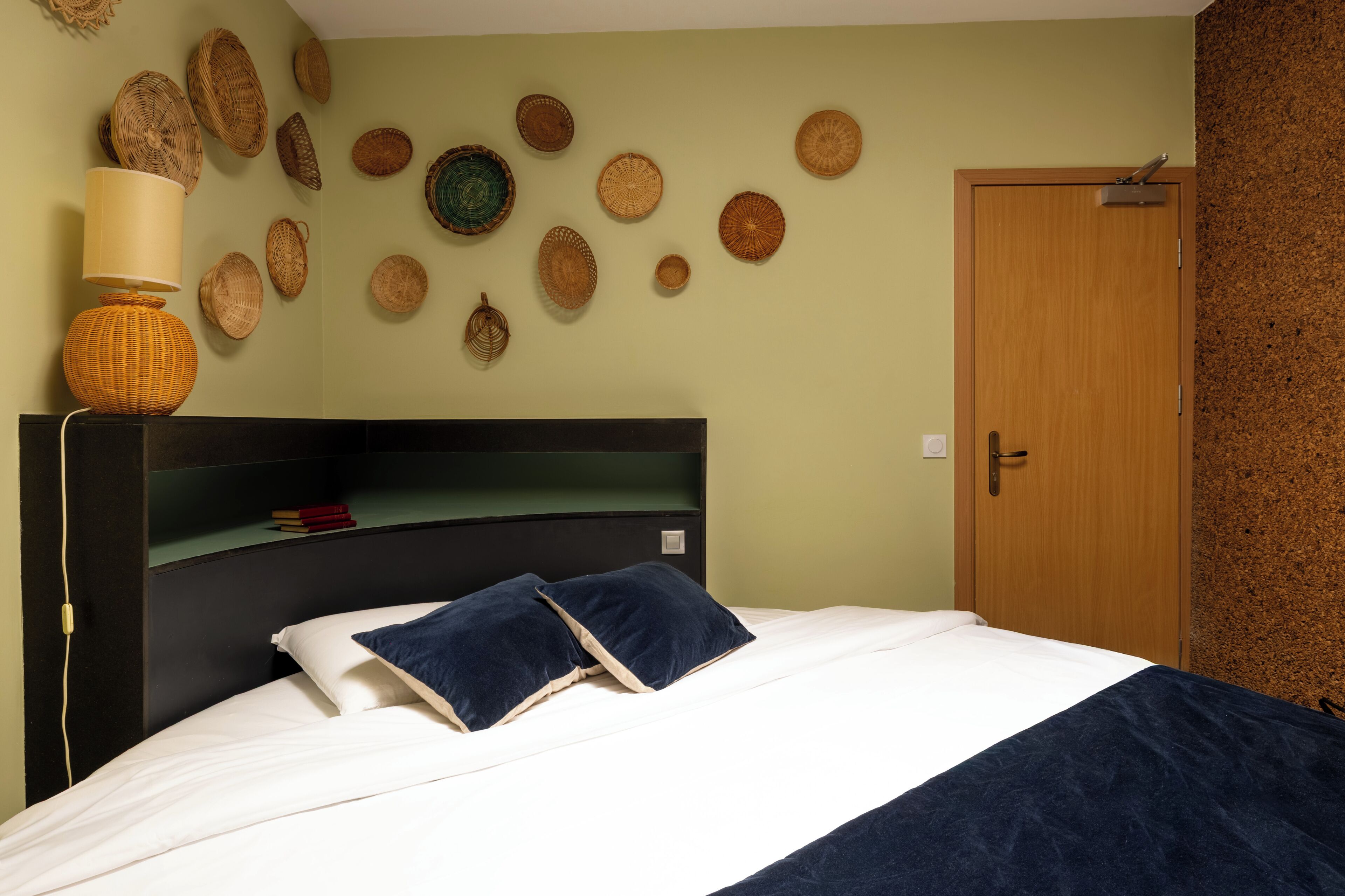 A bed with a dark blue bedspread and pillows, a bedside lamp, and a wall with woven baskets.