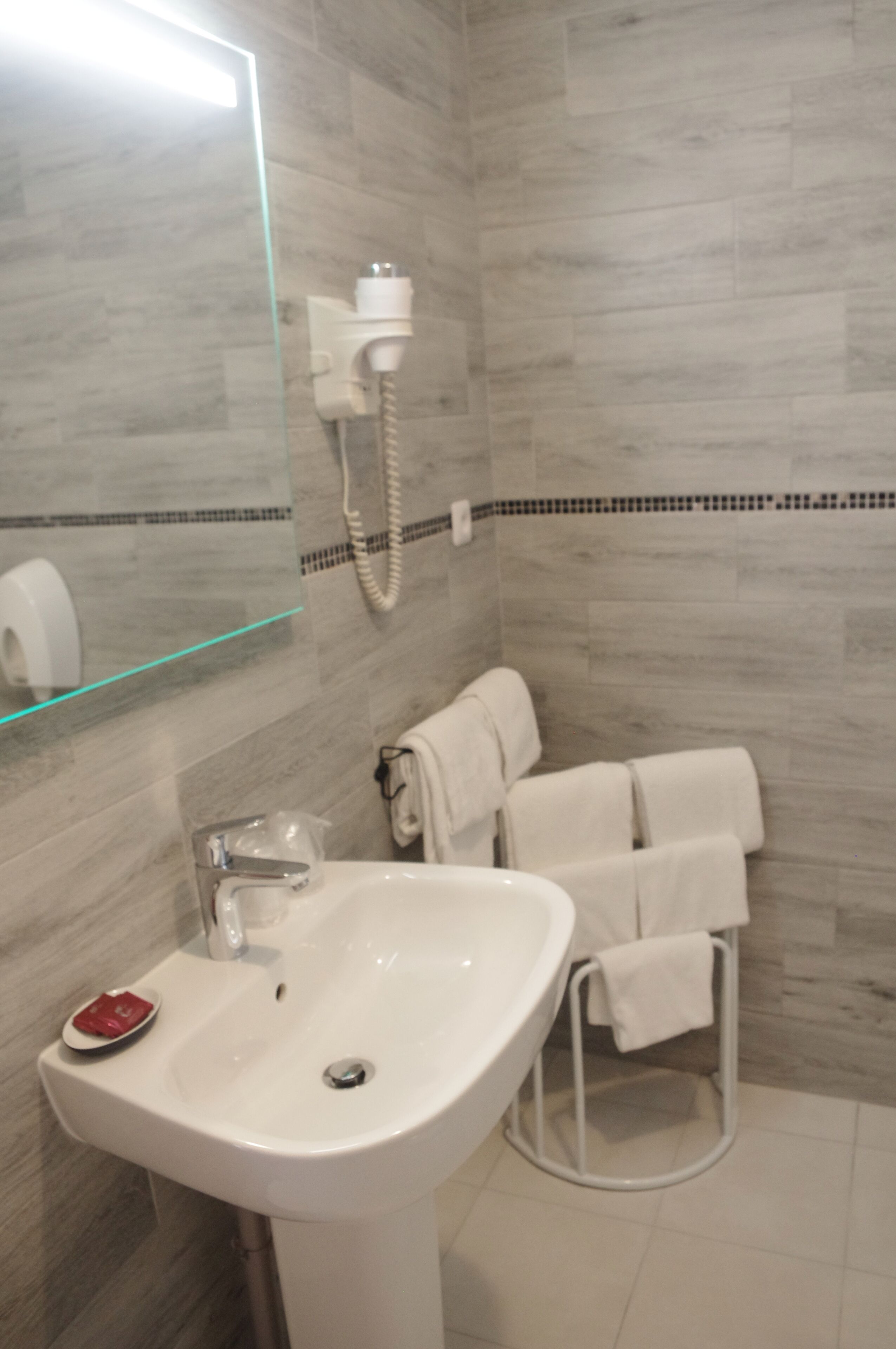 Une salle de bain avec un lavabo blanc, une cabine de douche en verre, un porte-serviettes avec des serviettes et un miroir avec un bandeau lumineux.