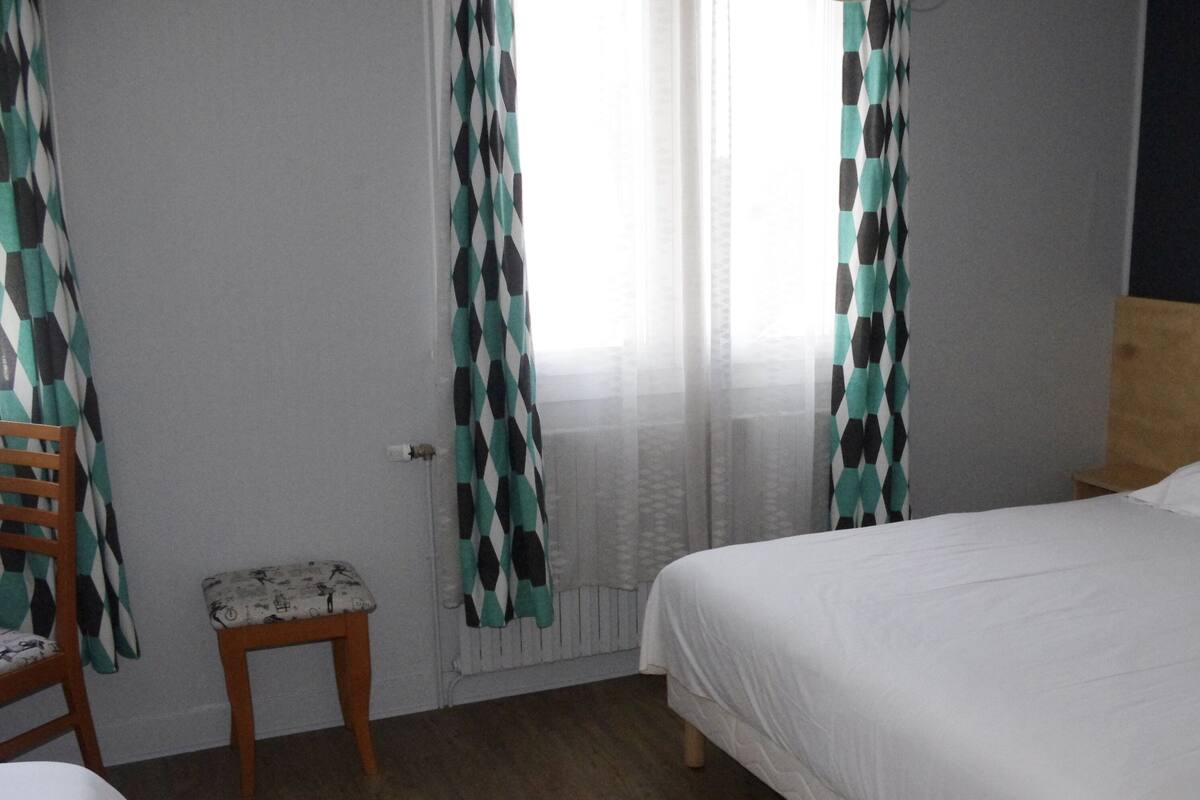 Un dormitorio con cama, cabecera de madera, mesita de noche, una silla y cortinas.