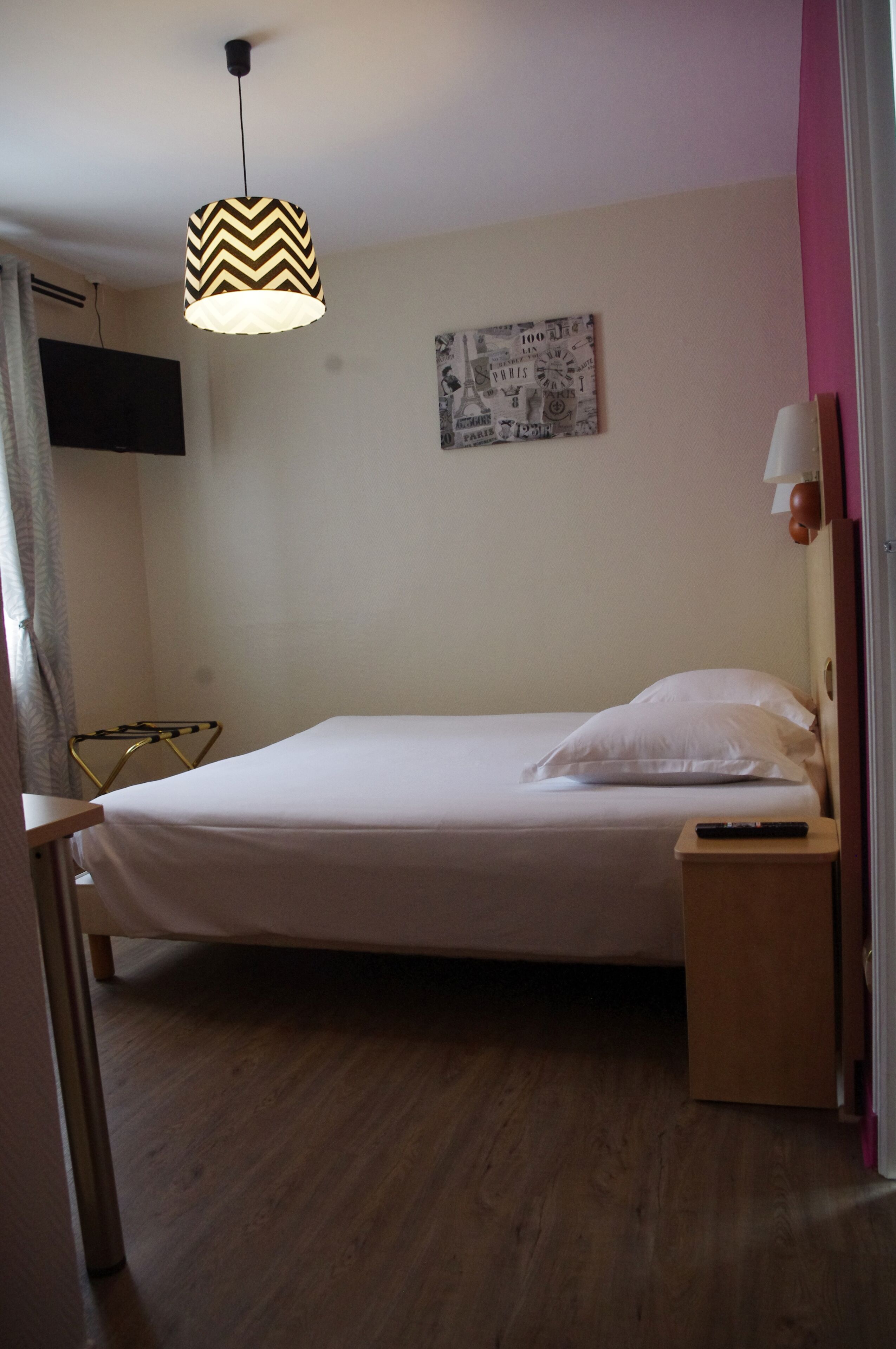 Una cama bien tendida con edredón blanco, una mesita de noche con lámpara, una lámpara colgante con diseño en zigzag, un reloj de pared y un televisor de pantalla plana montado en la pared.