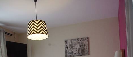 Una cama bien tendida con edredón blanco, una mesita de noche con lámpara, una lámpara colgante con diseño en zigzag, un reloj de pared y un televisor de pantalla plana montado en la pared.