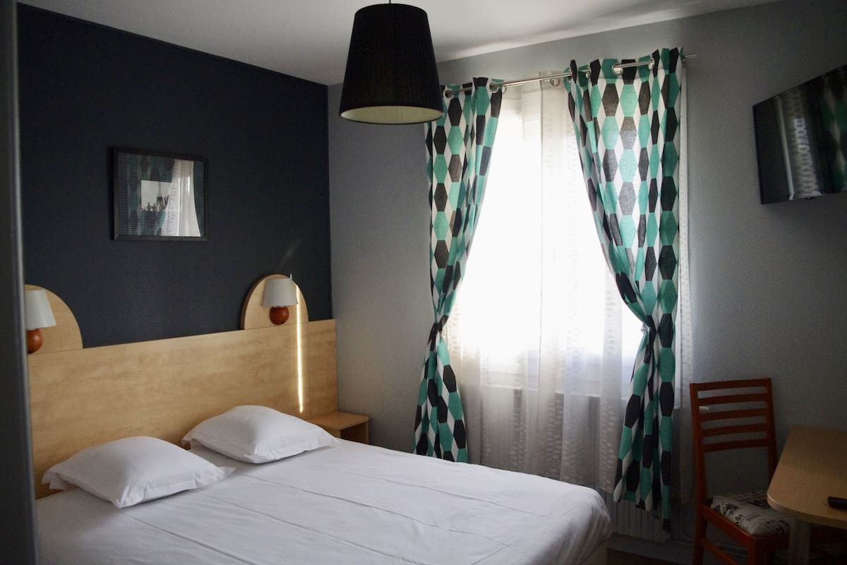 Una habitación de hotel con una cama, dos lámparas de noche, una silla, una mesa y un televisor en la pared.