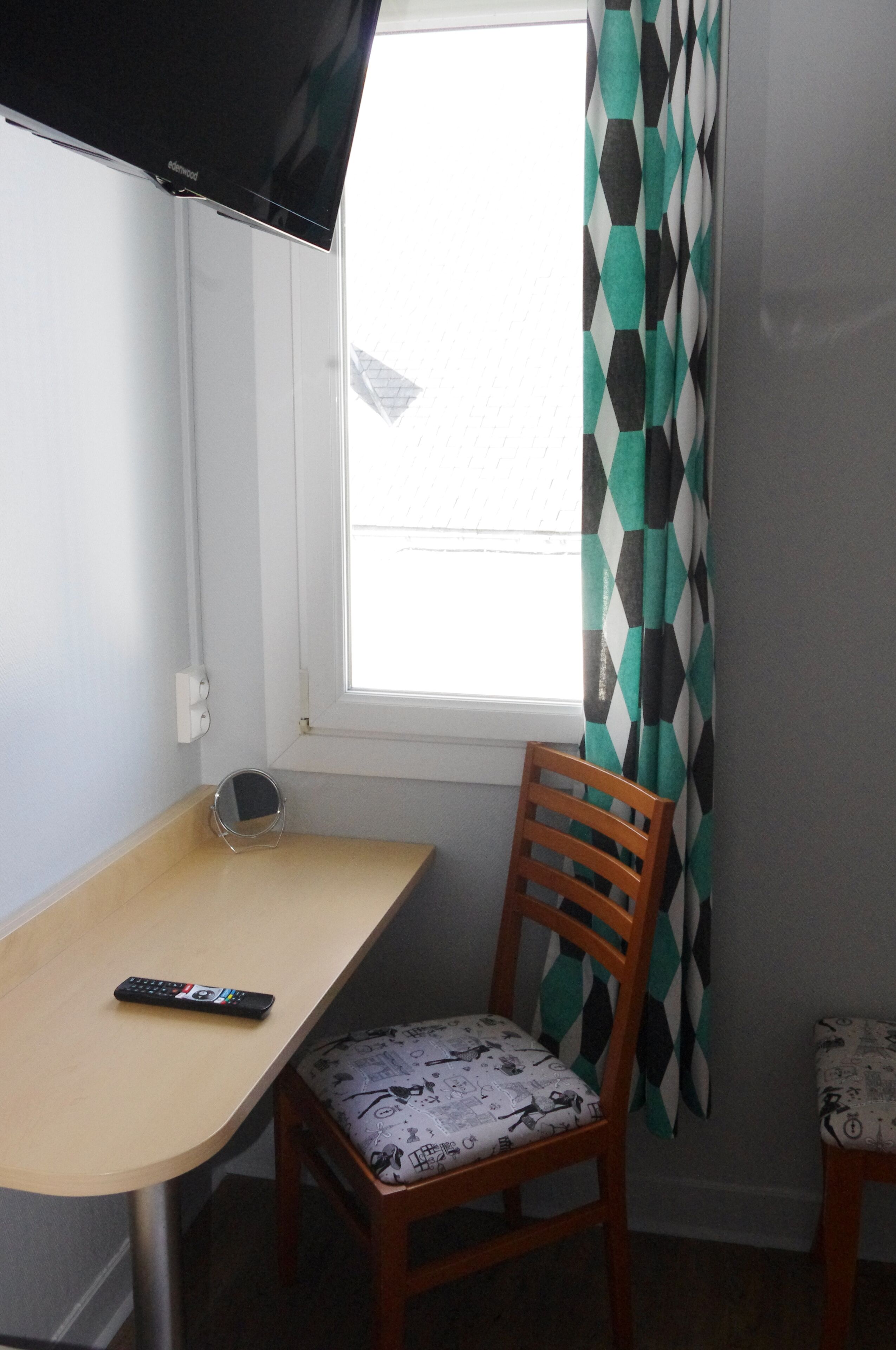 A small room with a window, a wooden chair with a patterned cushion, a small table, and a remote control on the table.