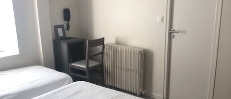 A bedroom with two beds, a radiator, a desk with a chair, a framed picture, and a door.