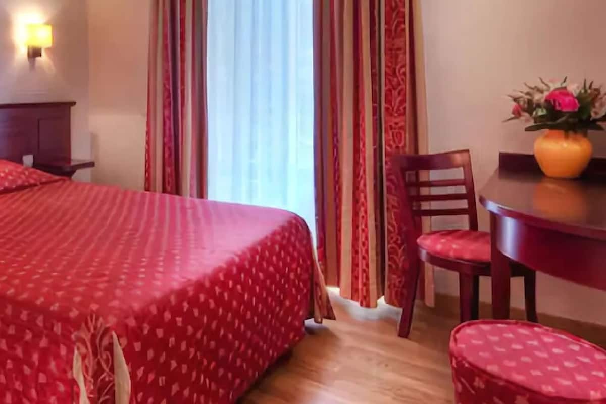 A hotel room with a large bed, a wooden nightstand, a desk with a chair, and a vase with flowers.