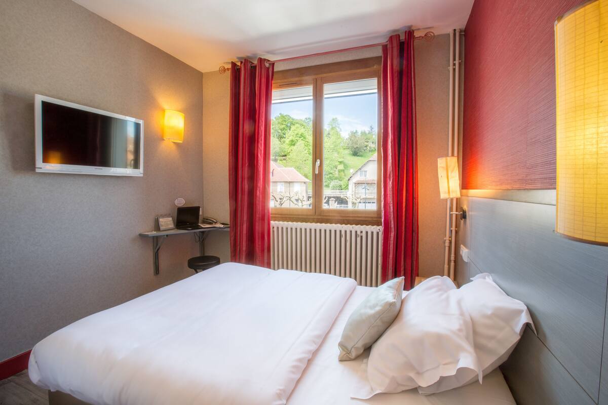 Ein Hotelzimmer mit Bett, Fernseher, Schreibtisch mit Computer und Fenster mit roten Vorhängen.