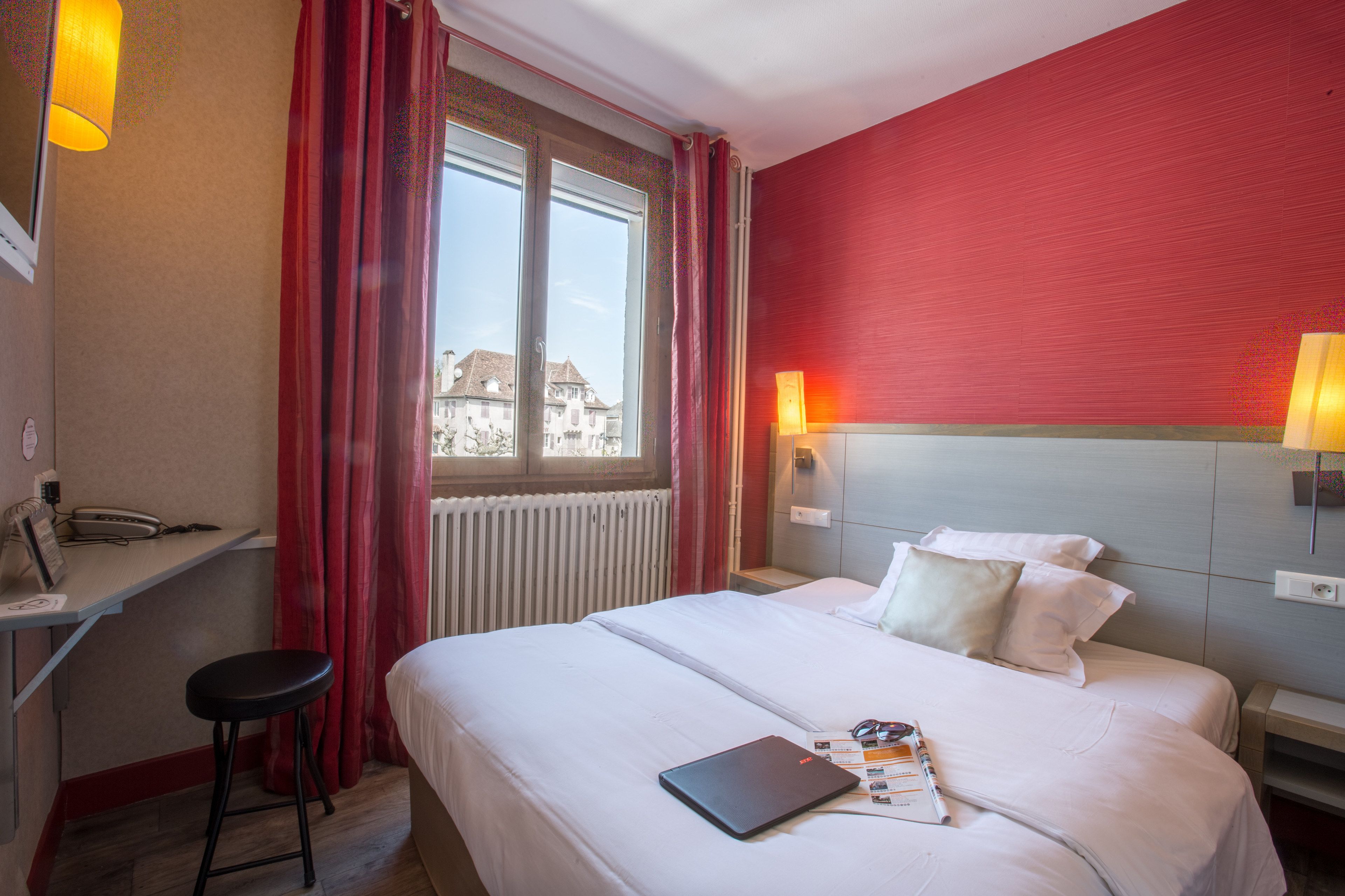 Habitación de hotel con cama, escritorio, silla, ventana con cortinas y vista a edificios.