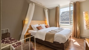 Ein Schlafzimmer mit einem großen Bett, einem Nachttisch, einem Hocker, einem Holzsessel und einem Fenster mit Vorhängen.