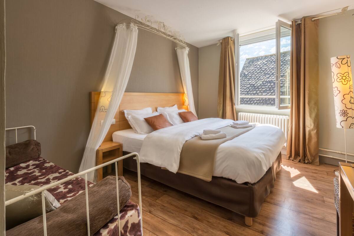 Ein Schlafzimmer mit einem großen Bett, einem Nachttisch, einem Hocker, einem Holzsessel und einem Fenster mit Vorhängen.