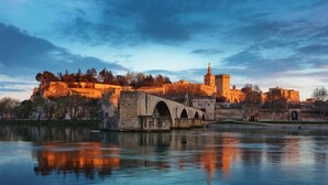 Een historische stenen brug over een rustige rivier, leidend naar een versterkt kasteel met een opvallende toren.